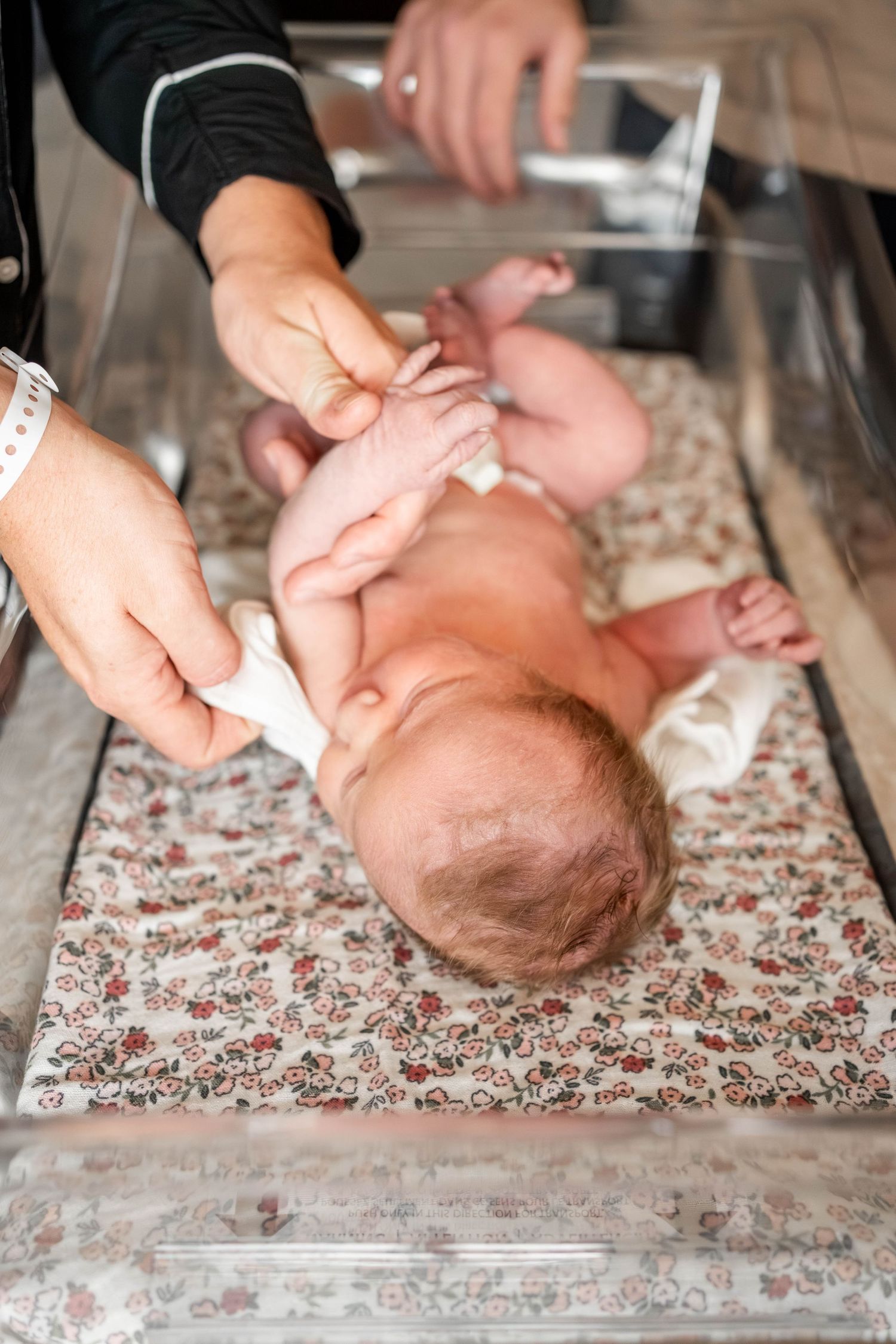 Newborn baby lying on floral mat while caregiver gently holds legs during diaper change in hospital nursery.