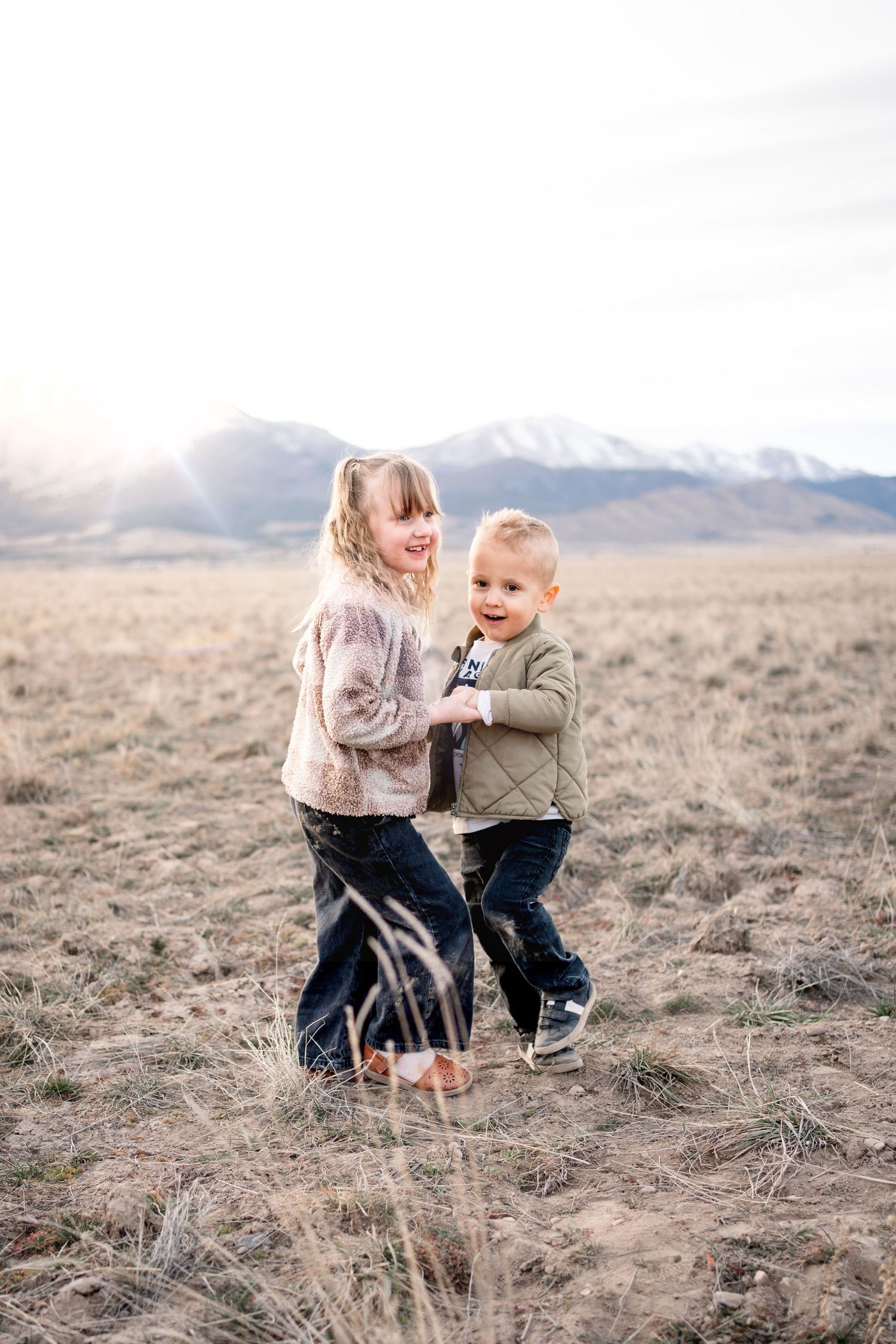 Two people share a playful moment in a field with snow-capped mountains visible in the background.