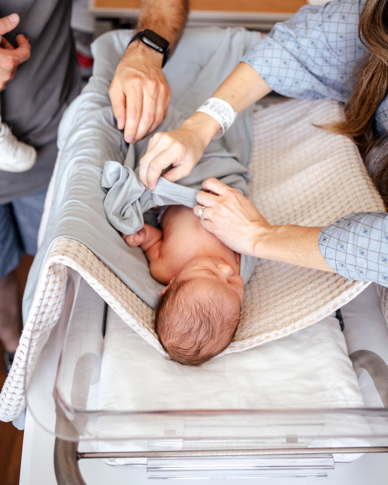 Newborn baby lying in hospital bassinet while parents in medical setting gently adjust swaddle blanket after birth.