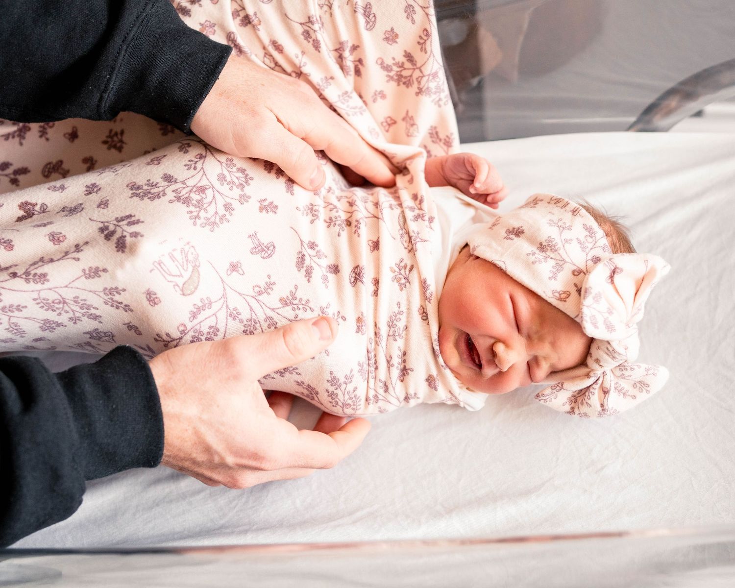 Newborn baby in floral outfit and matching bow headband crying on changing table held by parents.