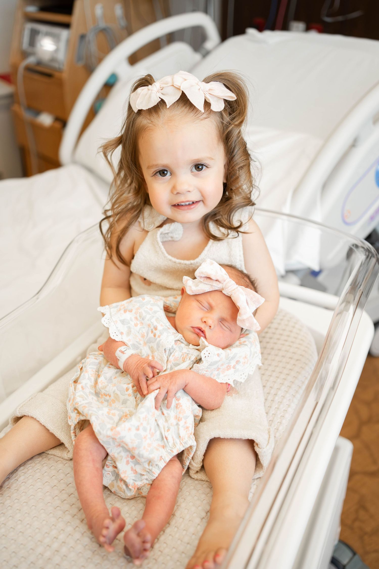 Young girl in beige dress holds sleeping newborn baby sibling in hospital bassinet, both wearing floral outfits.