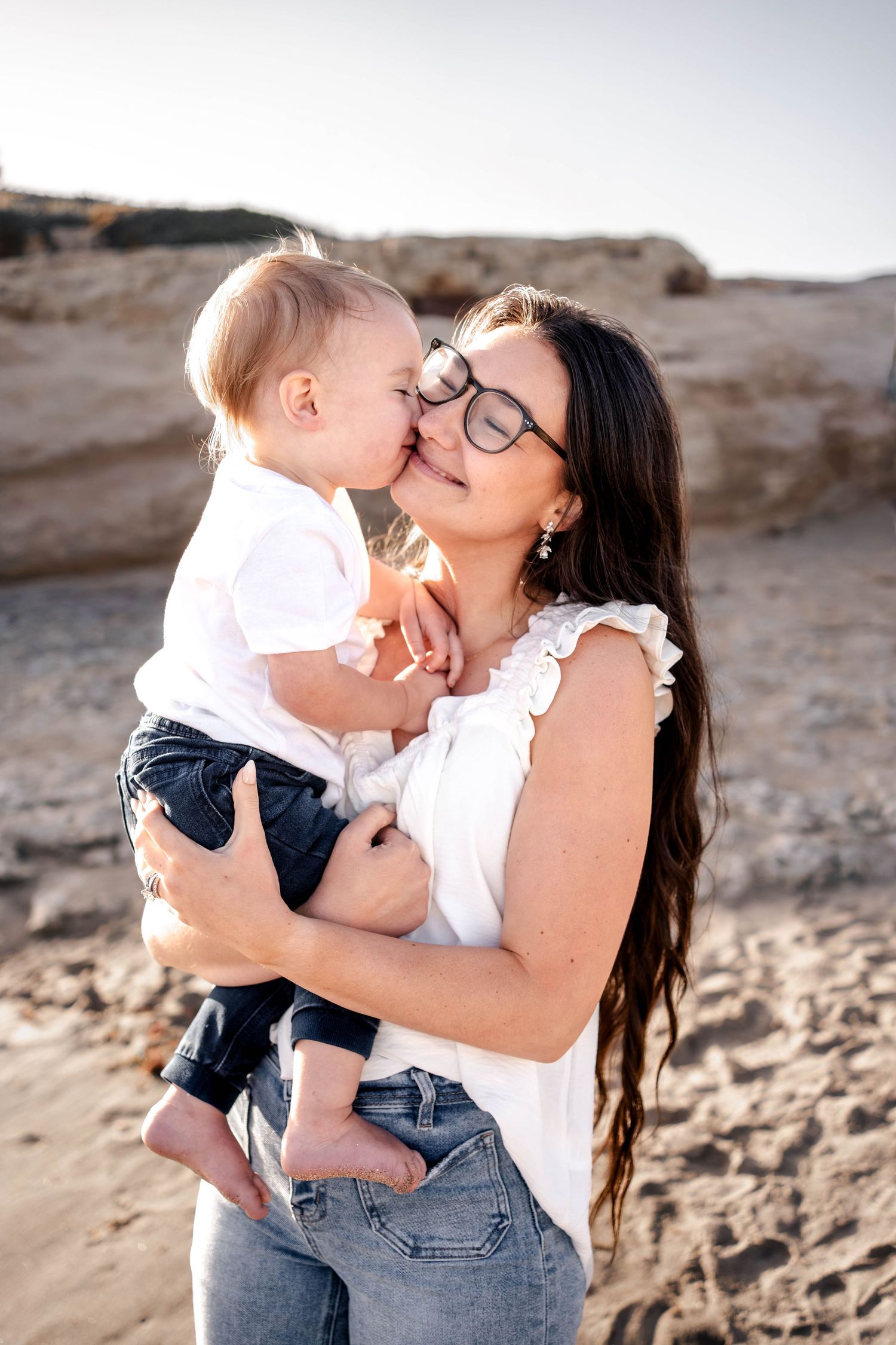 A mother with dark hair and glasses holds her baby on a sandy beach, sharing a tender nose kiss outdoors.