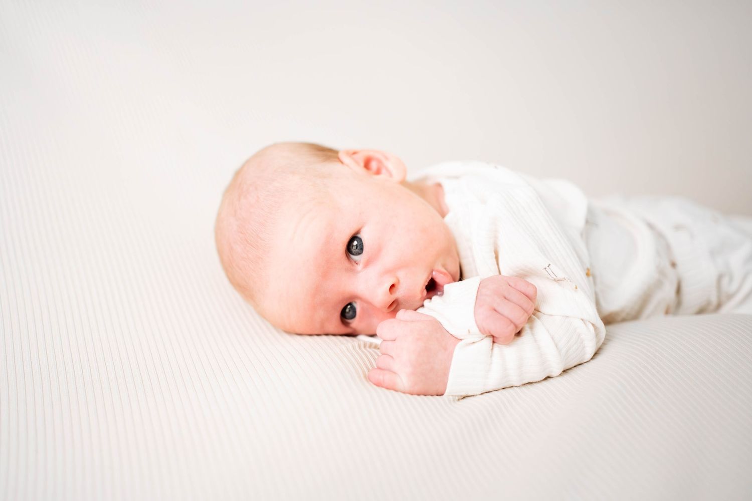 Newborn baby in white onesie lying on a cream textured surface, looking at the camera with bright eyes.