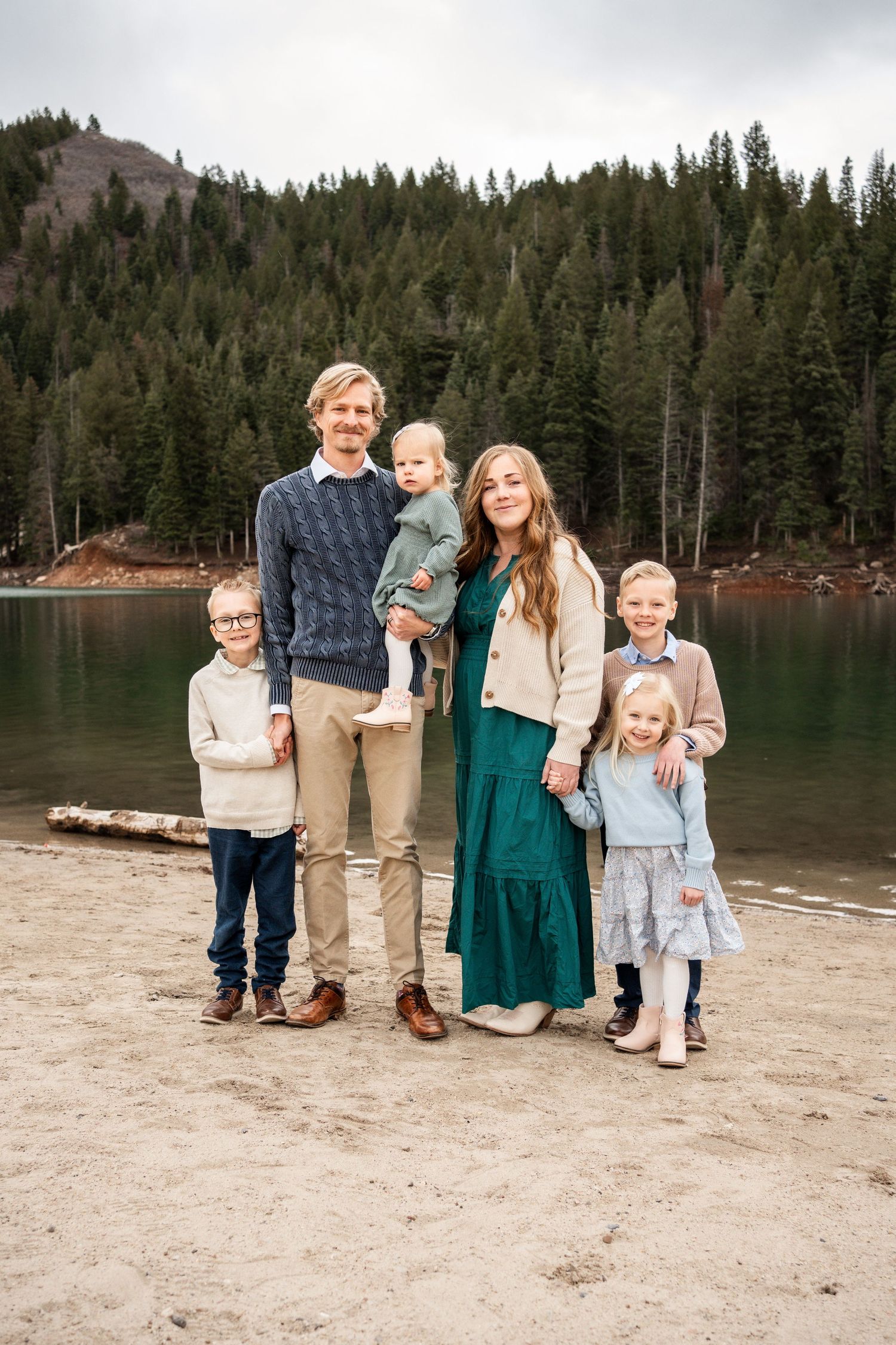 Family in coordinated neutral outfits poses together on sandy beach with evergreen forest and mountain backdrop.