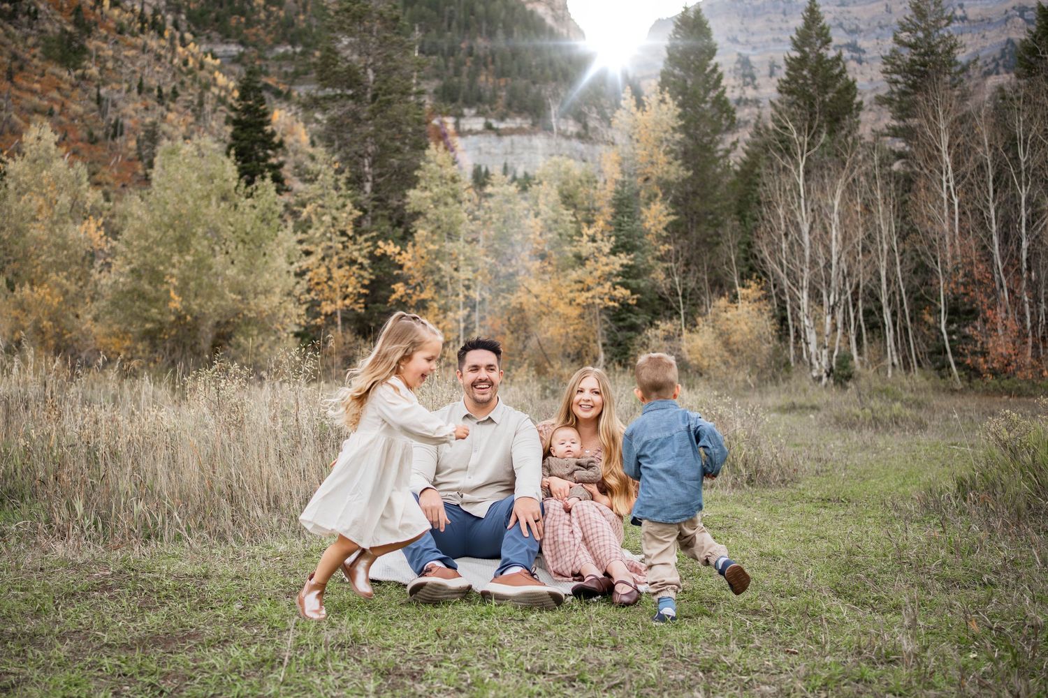 Family of five sitting together on a blanket in a grassy mountain meadow surrounded by golden autumn trees at sunset.