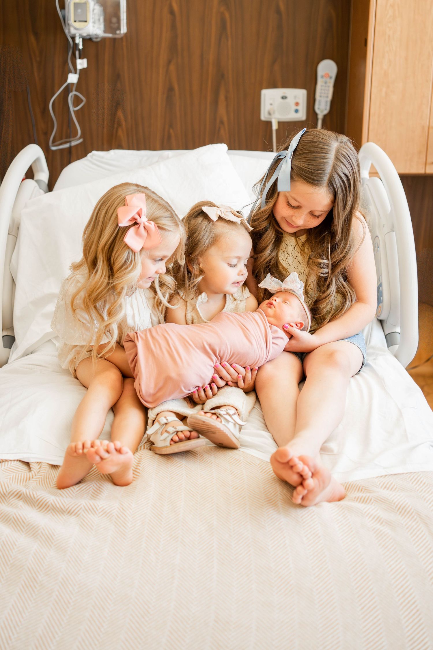 Three young girls sit on a hospital bed, lovingly gazing at their newborn sibling wrapped in a pink swaddle.