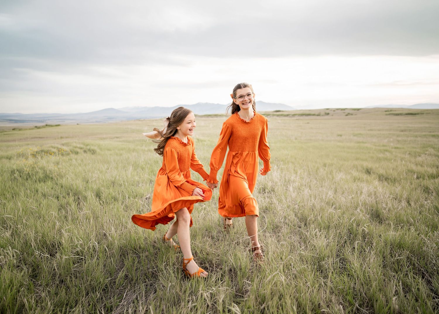 Two girls in matching orange dresses hold hands running through a green field with mountains in the background.