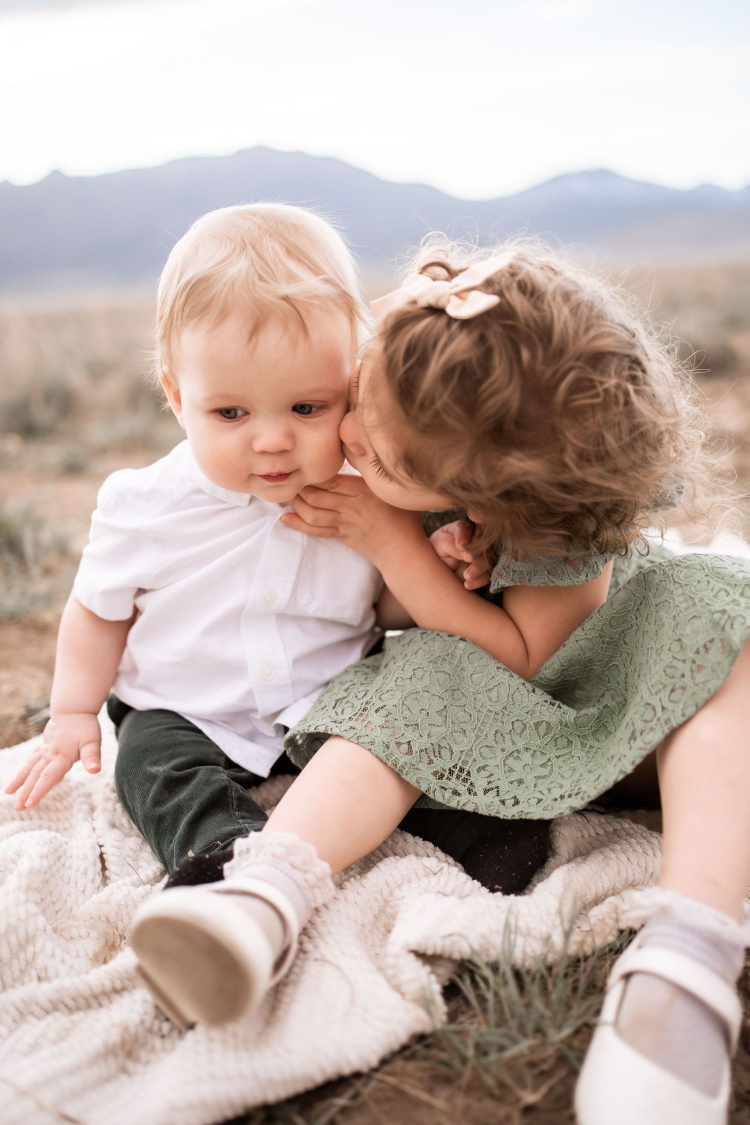 A toddler girl in a green lace dress gently kisses a baby boy on a blanket with mountains in the background.