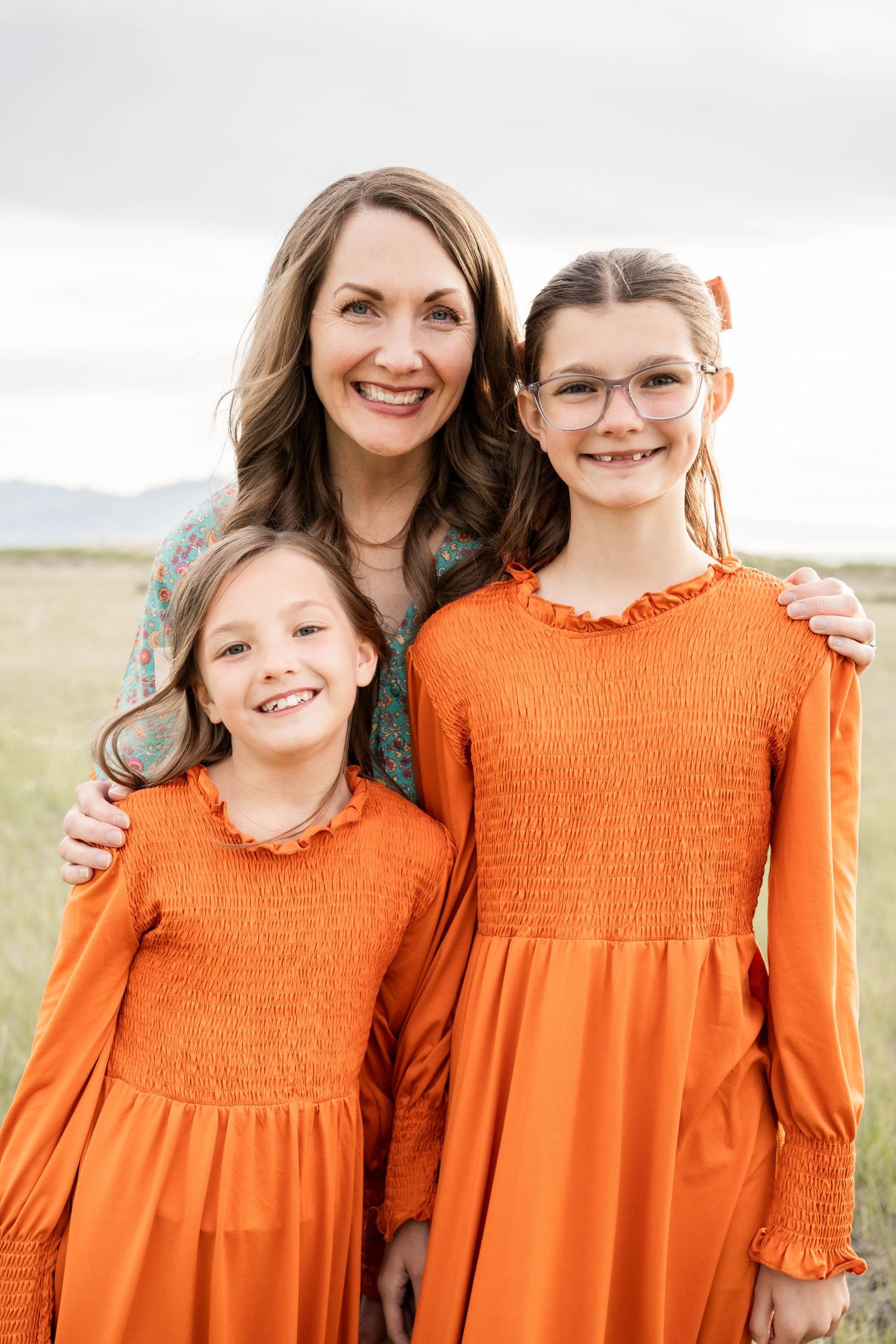 A woman and two girls in matching orange dresses smile together outdoors in a field.