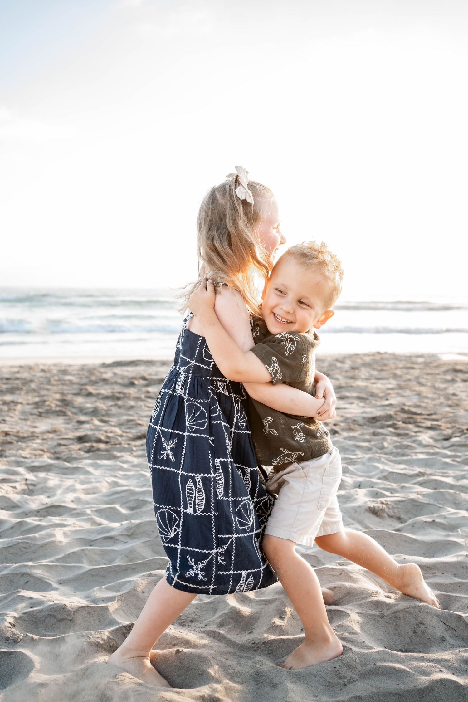 Two young children hugging and playing on a sandy beach, wearing navy and olive outfits, with ocean waves in the background.