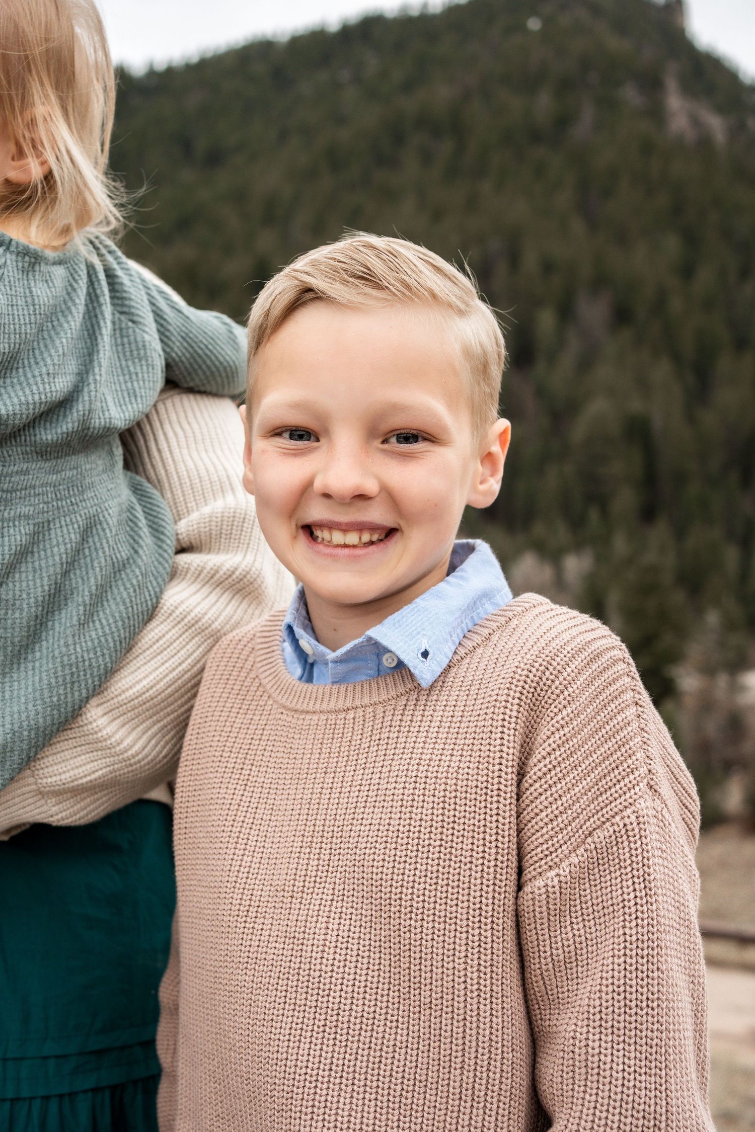 A young person wearing a beige knit sweater over a collared shirt stands outdoors with mountains in the background.