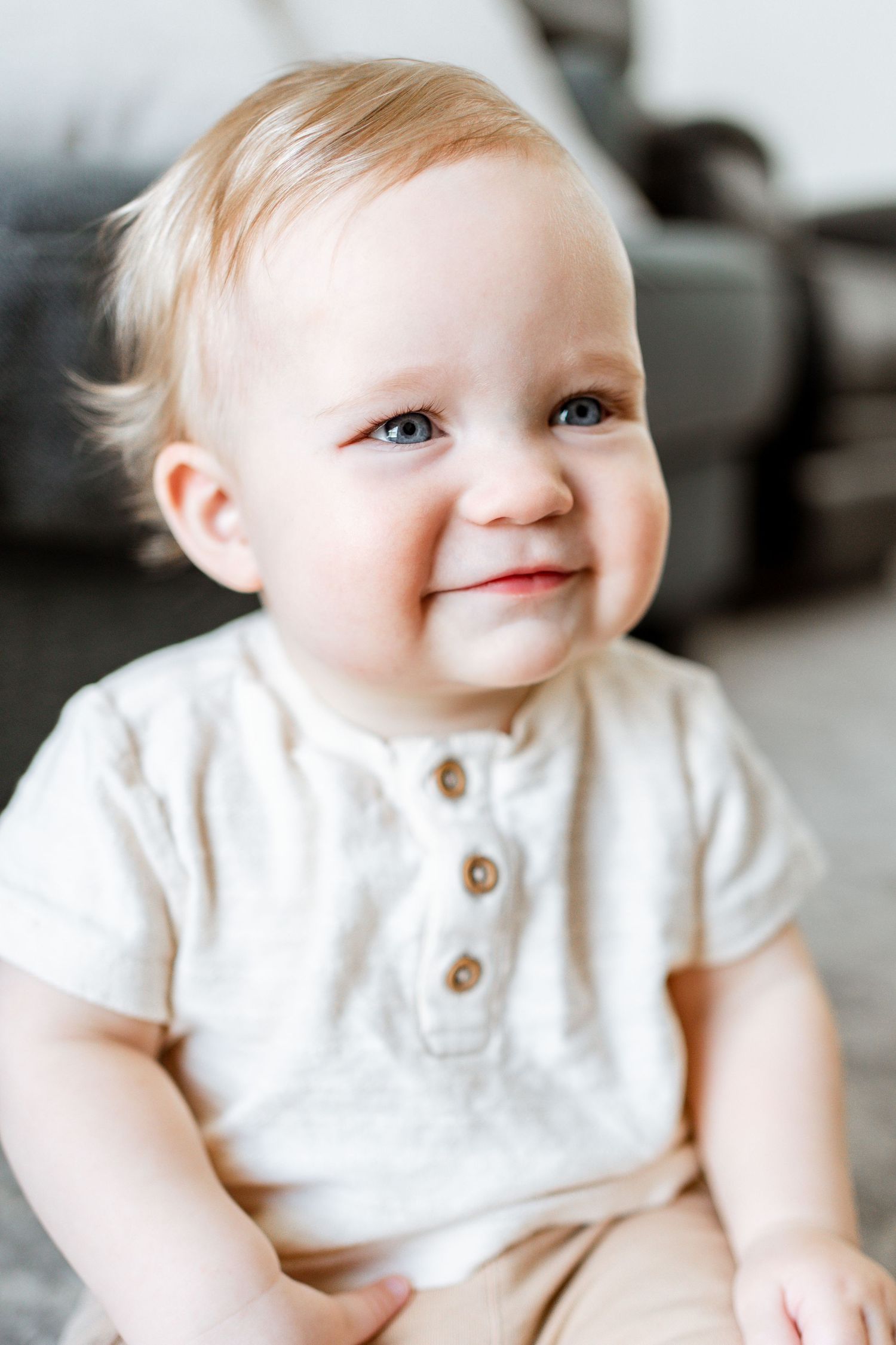 Infant in white button-up outfit smiles while sitting against light background.