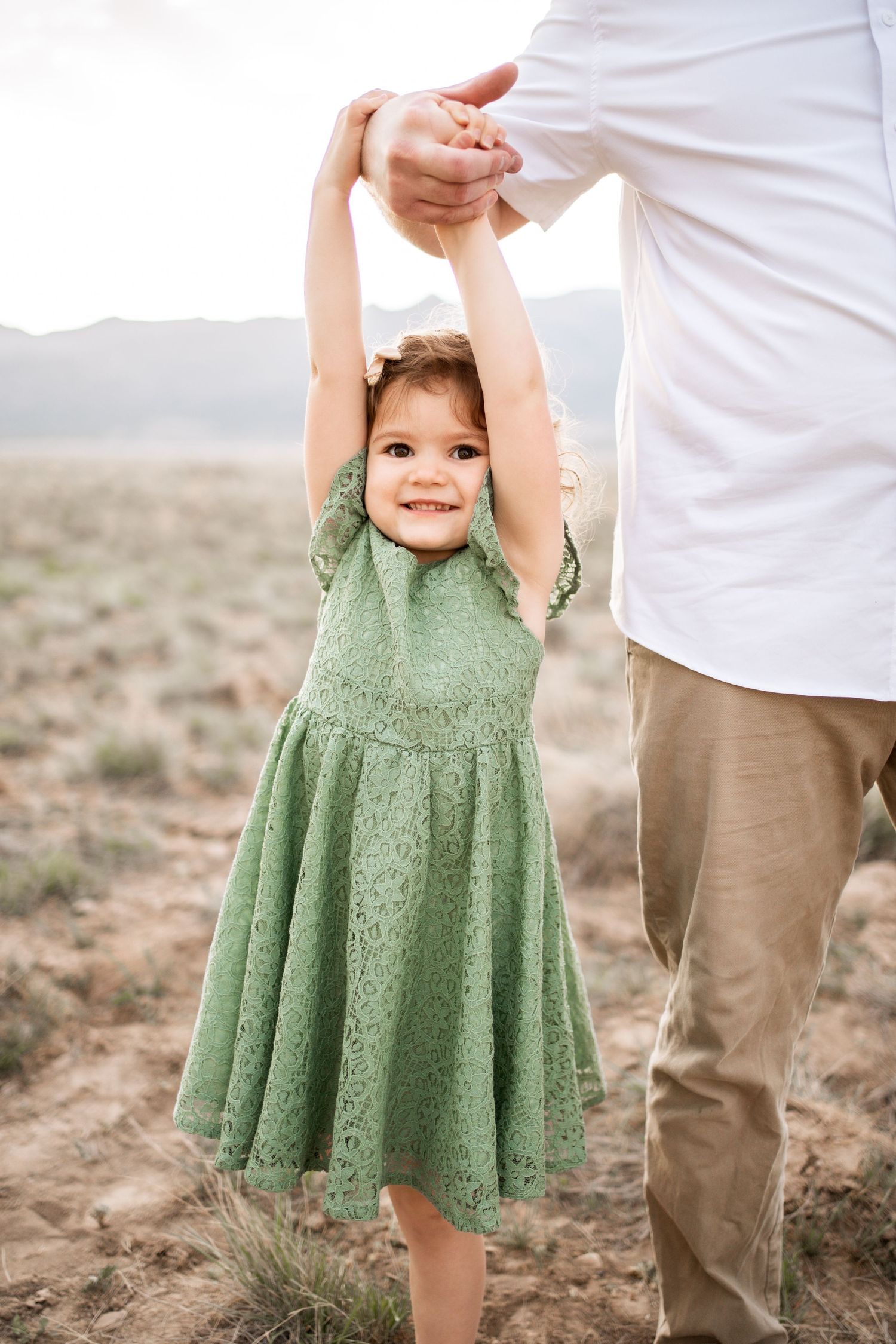Happy toddler girl in green lace dress holds fathers hands up in sunny desert field.