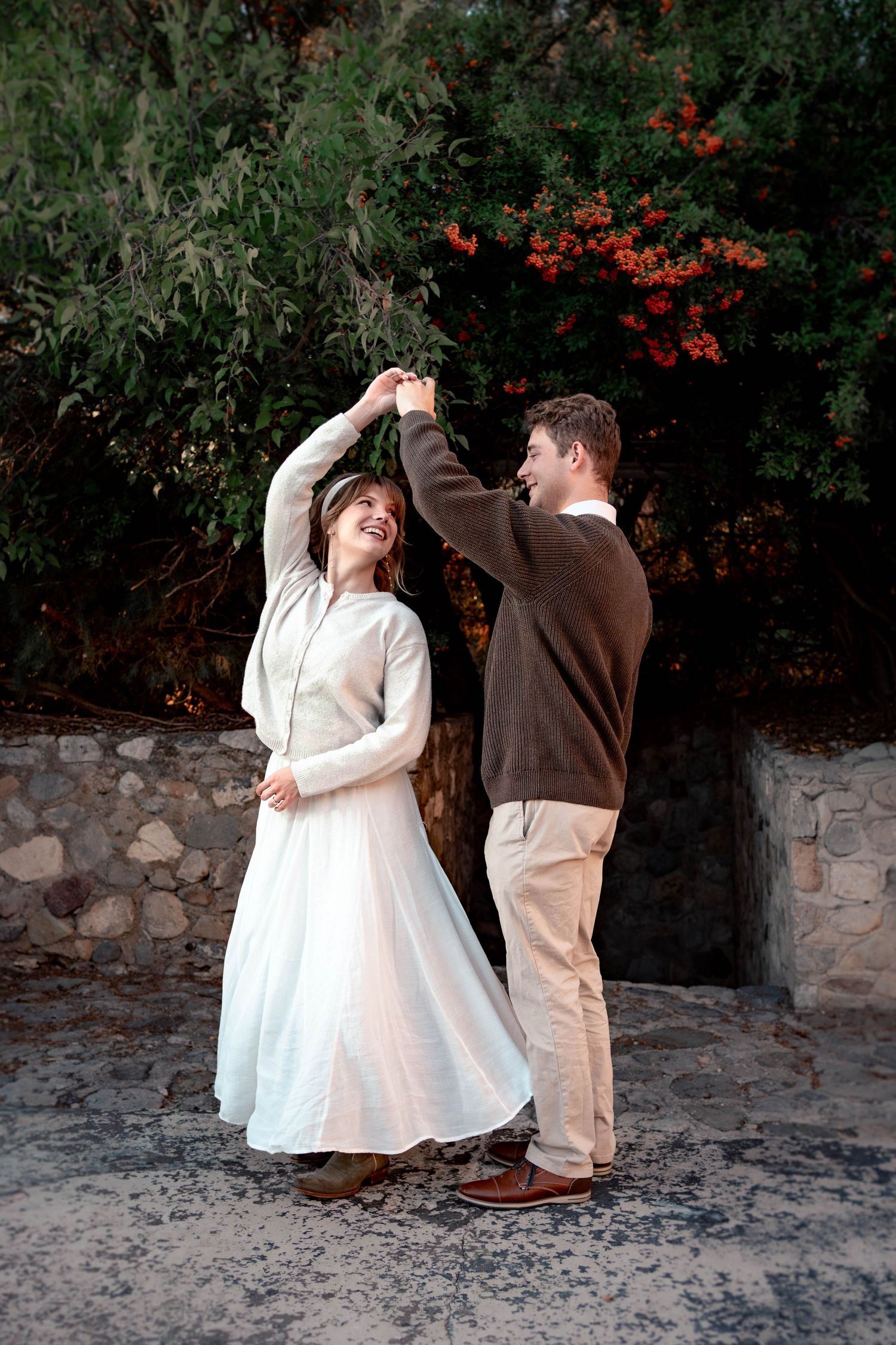 A smiling couple dances outdoors, the woman twirling in a white dress beside a stone wall and greenery with red flowers.