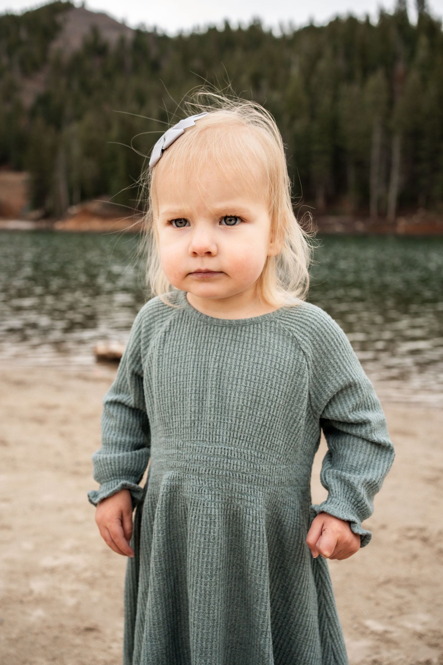 A small child in a sage green knit sweater stands on a beach near a lake with mountains in the background.