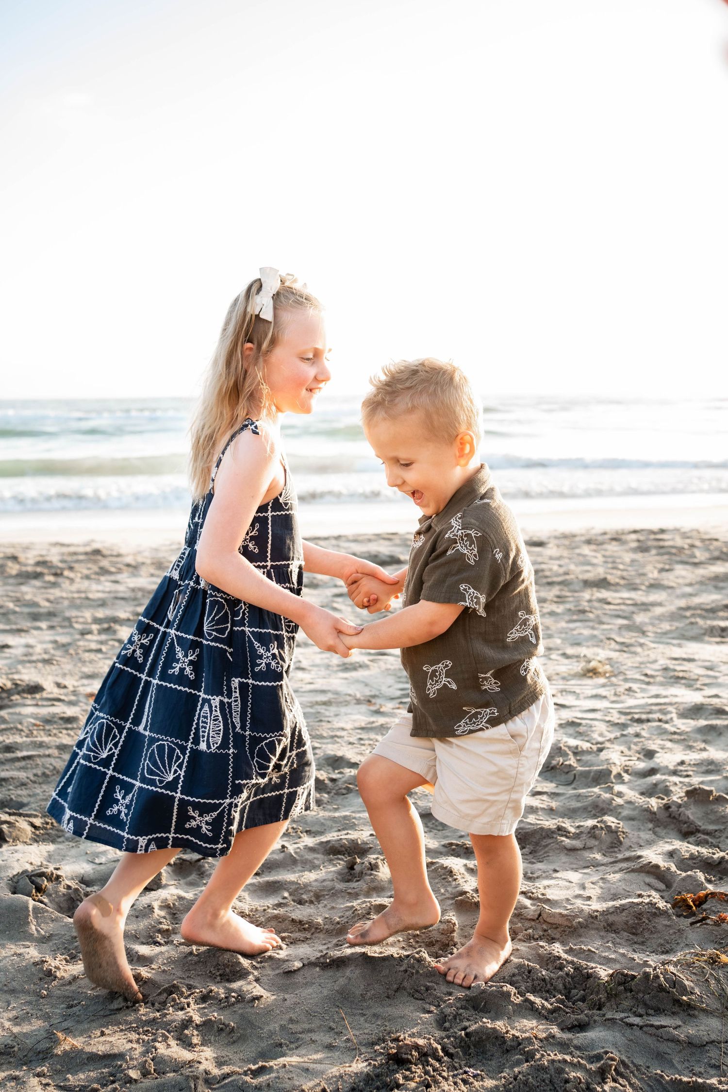 Two young children holding hands and playing on a sandy beach near the ocean on a sunny day.