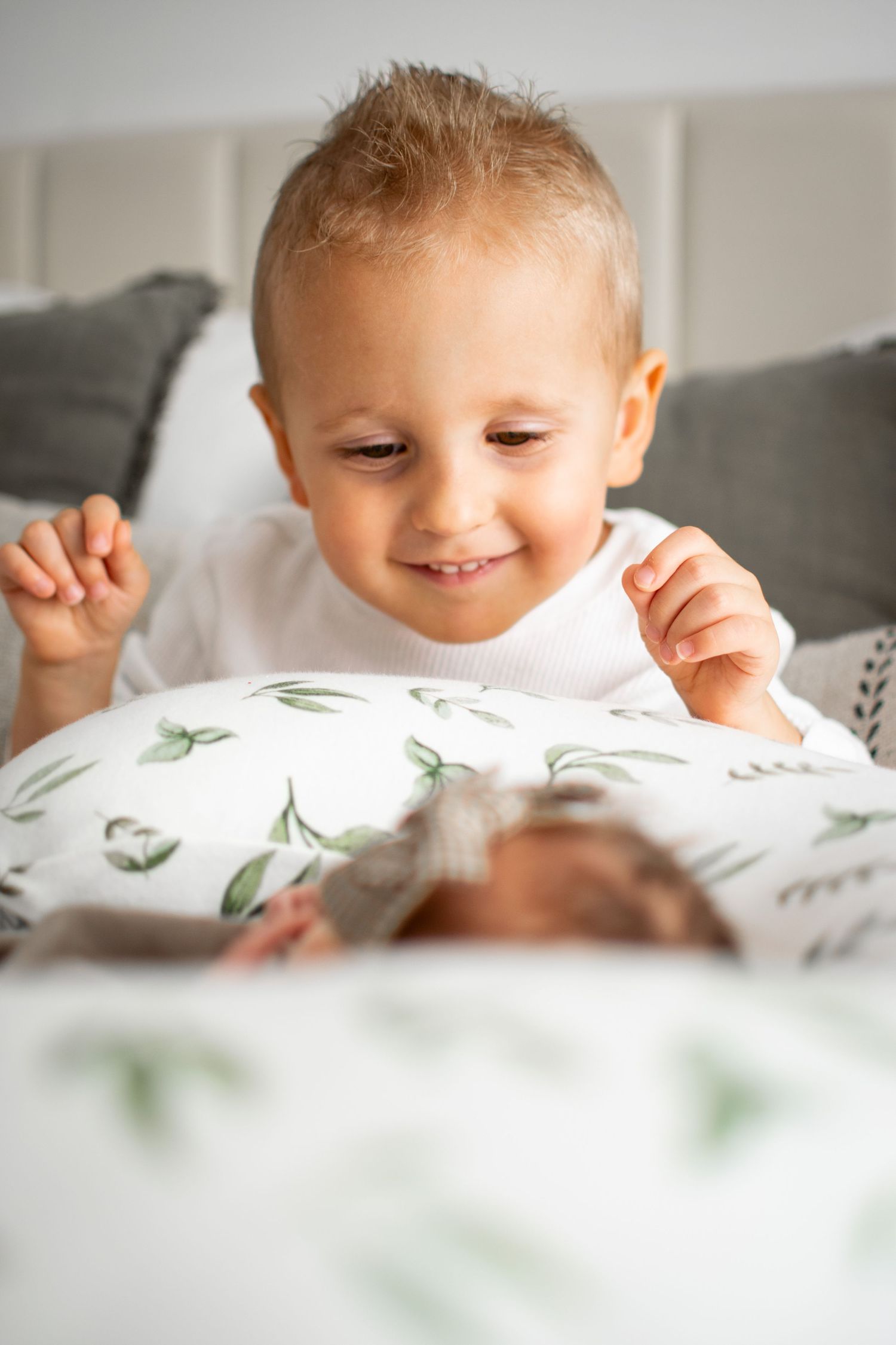 Child peeks over white pillow with green leaf pattern while playing peek-a-boo.