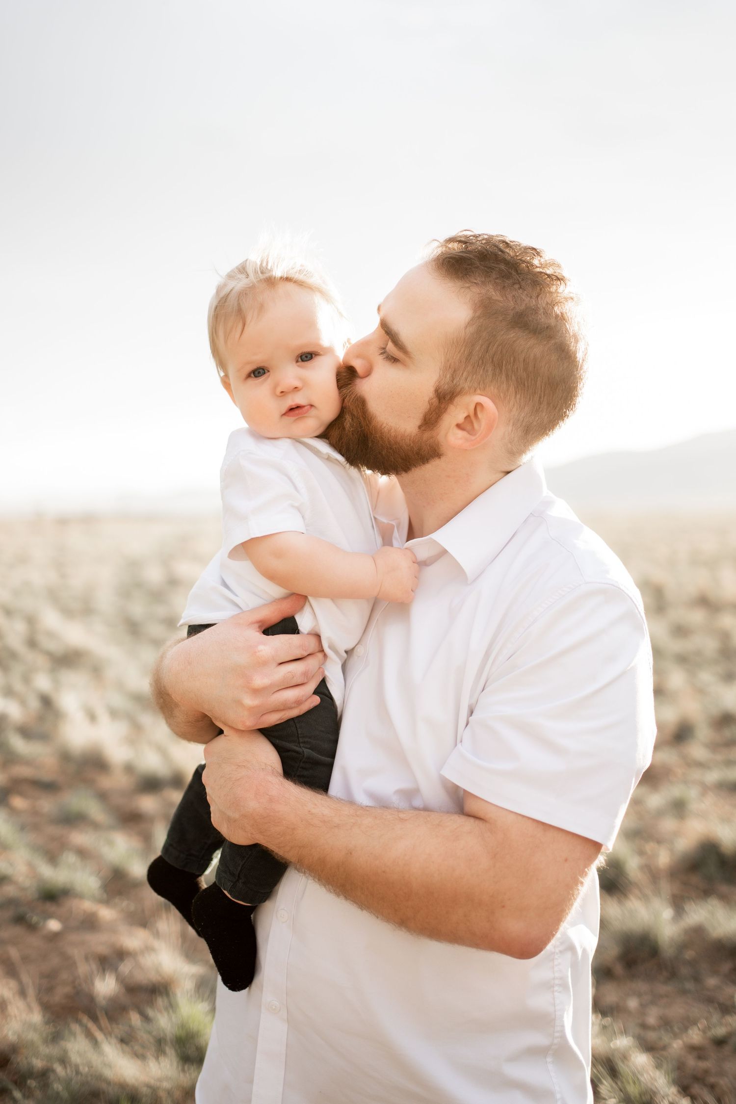 Bearded father kissing his baby boy outdoors in a desert field, both wearing white shirts.