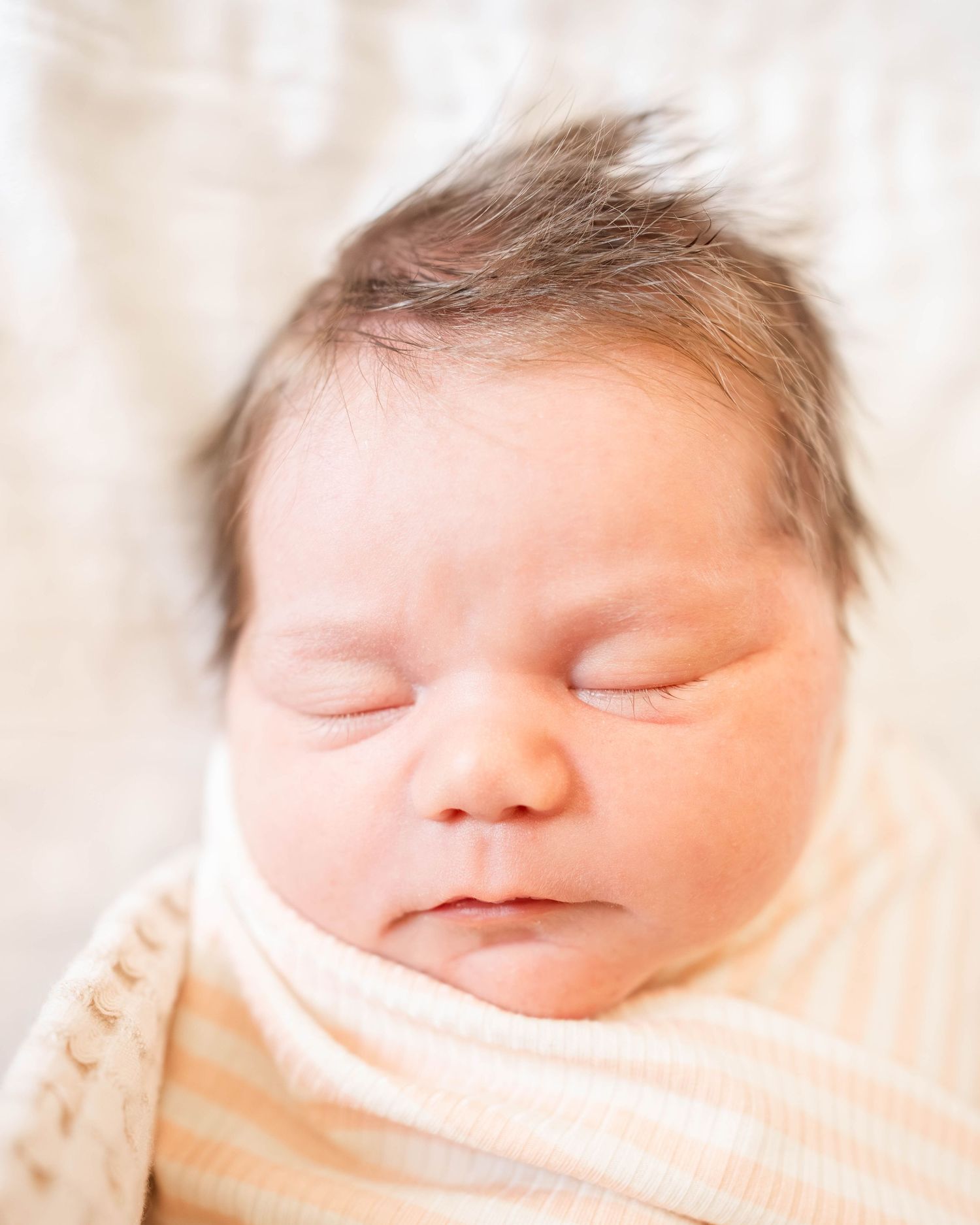 Peaceful sleeping newborn baby swaddled in a soft striped blanket with dark hair, captured in a close-up portrait.