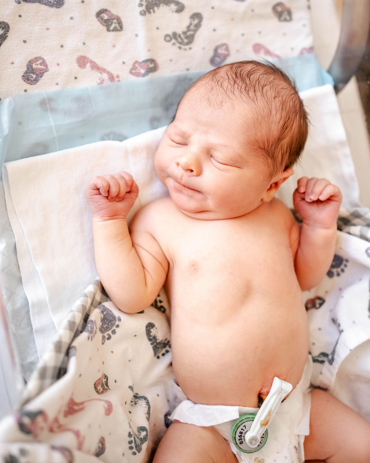 Peaceful newborn baby sleeping with fists raised, wearing a diaper, lying on a patterned blanket in a hospital setting.