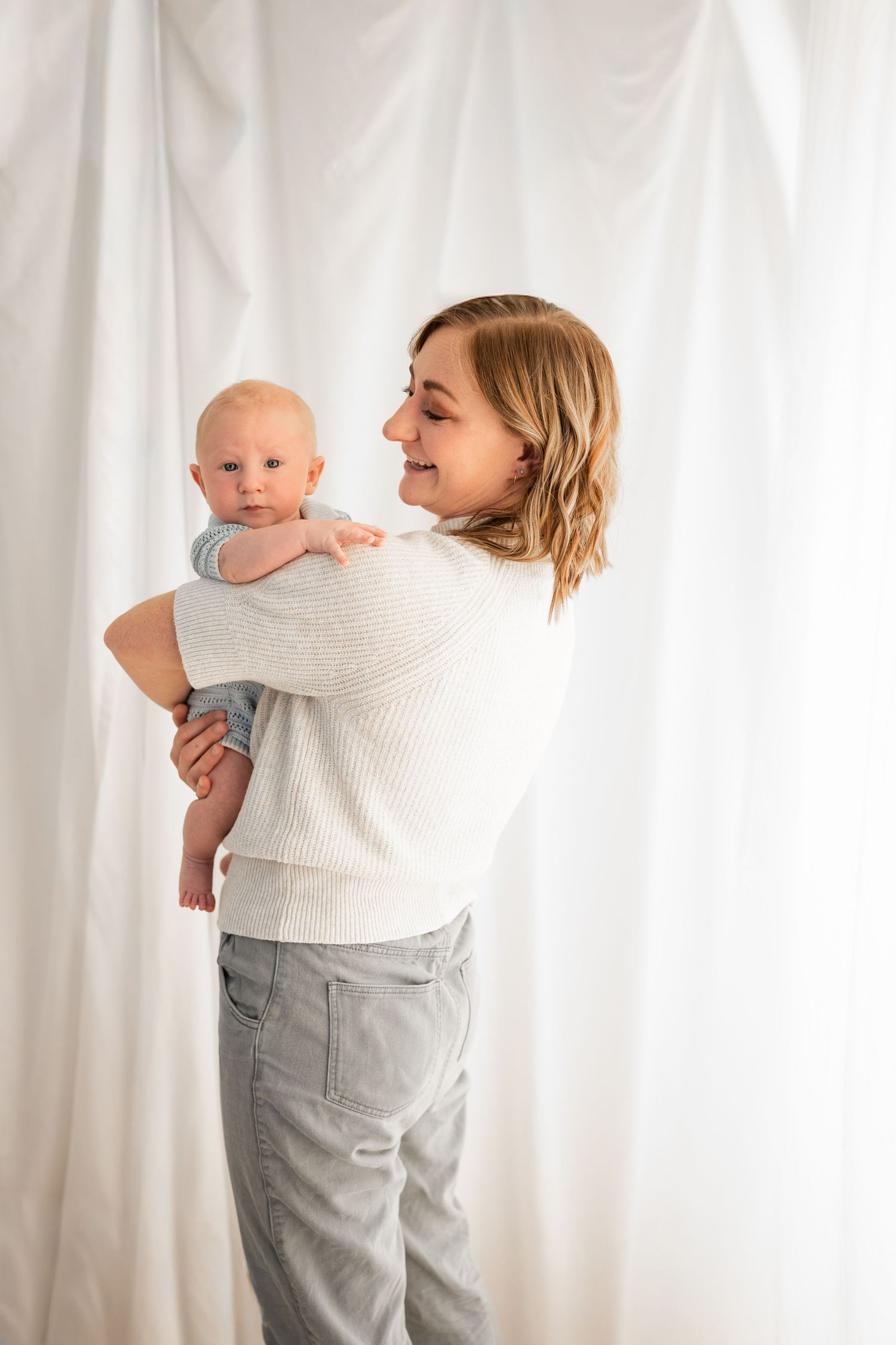Smiling woman in a cream knit sweater and gray pants holds a baby over her shoulder against a soft white draped backdrop.