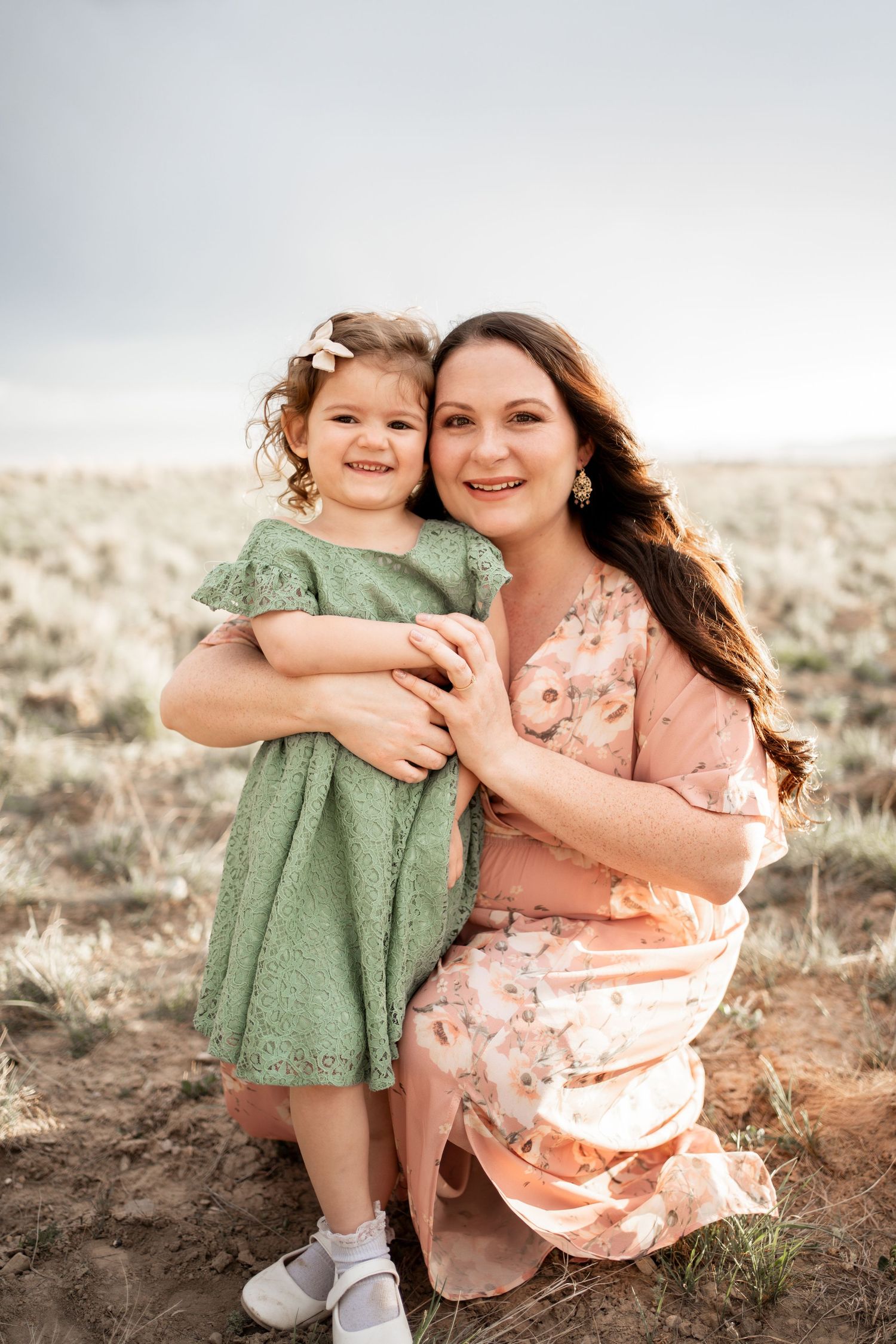 A woman in a floral dress crouches outdoors hugging a smiling toddler girl in a green dress in an open field.