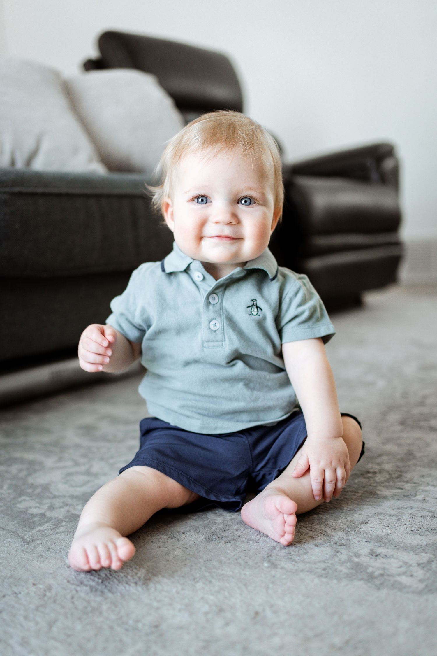 Baby wearing light blue polo and navy shorts smiles while sitting on gray carpet by dark couch.