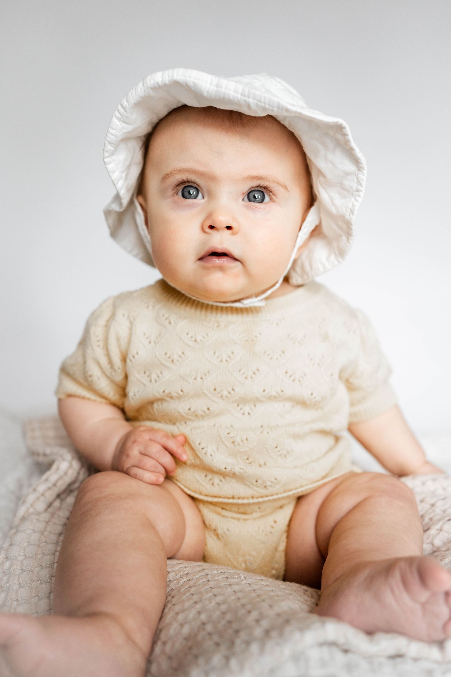 Adorable baby in cream knit romper and white sun hat sitting upright on a soft blanket.