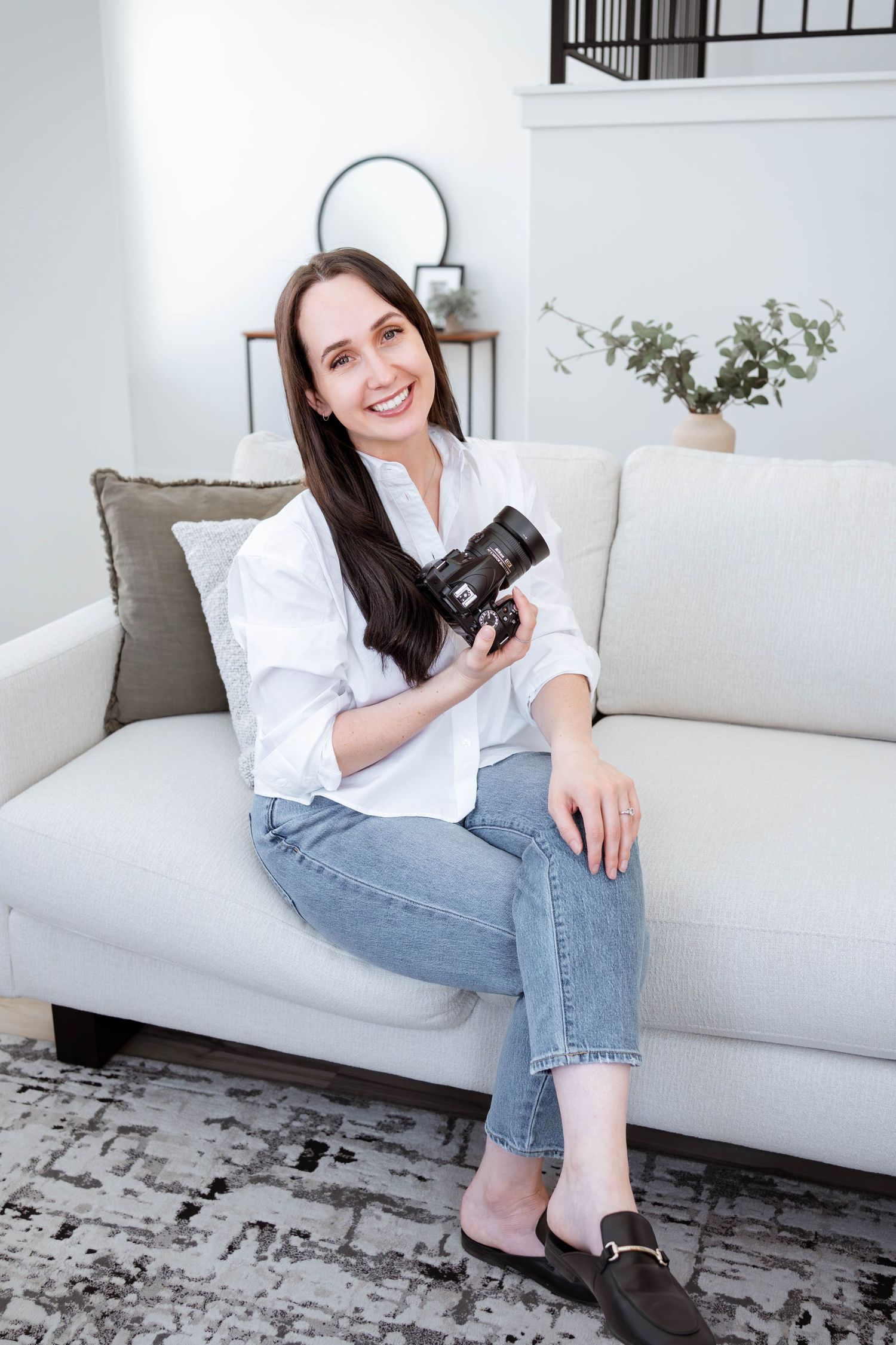 Smiling woman with dark hair sitting on white sofa holding a camera, wearing white shirt and jeans in bright living room.