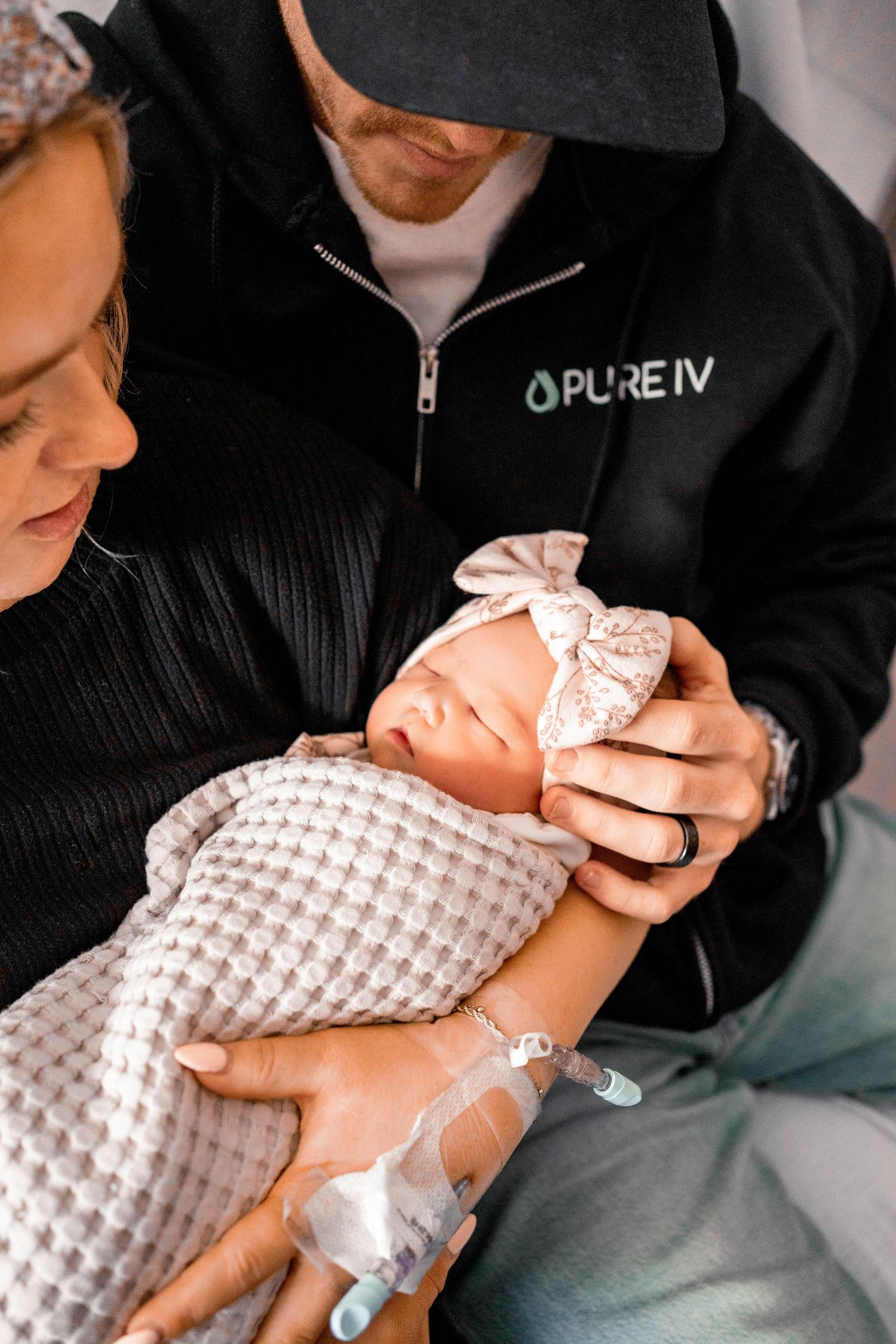 New parents cradle their sleeping newborn girl with a bow headband in a hospital room.