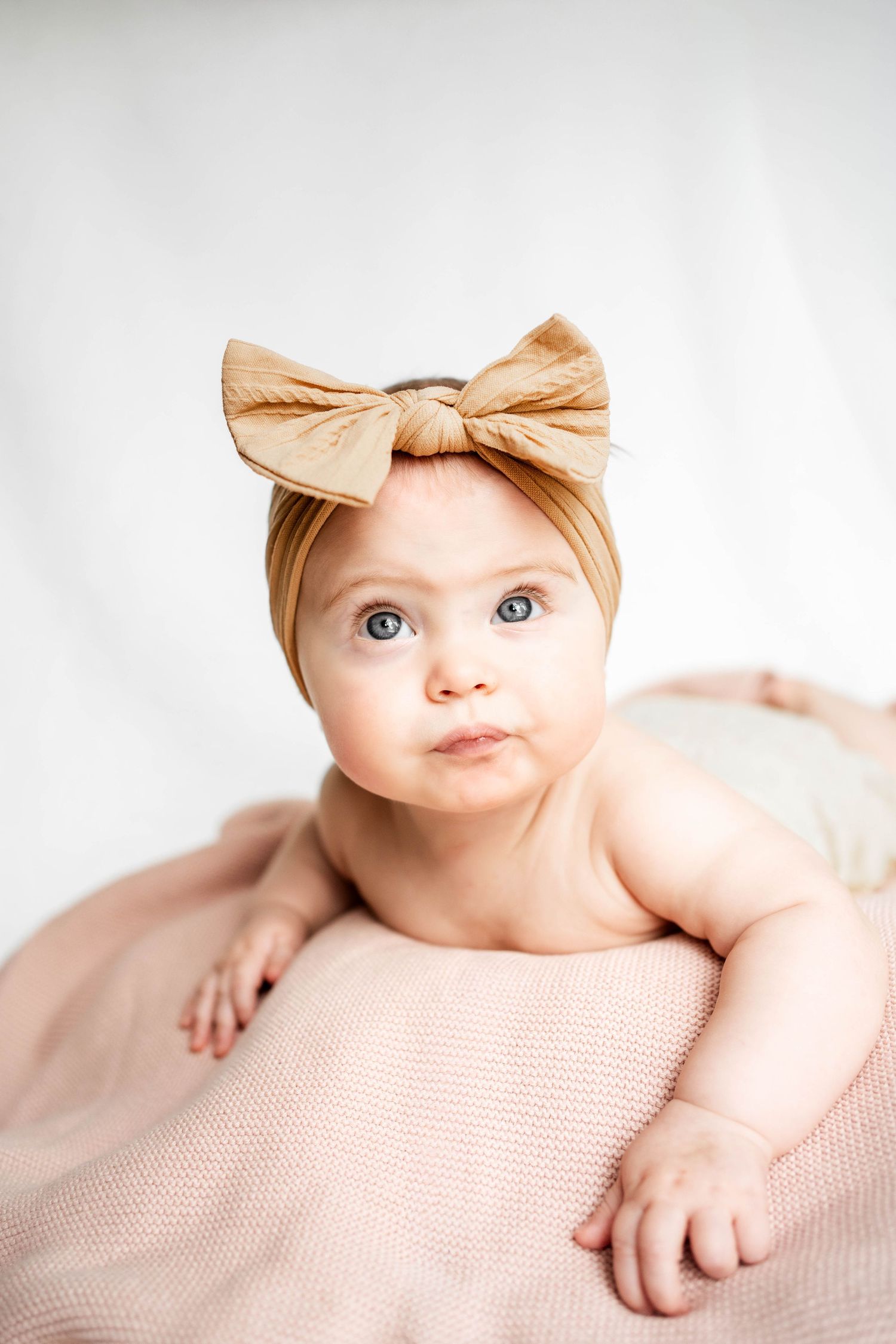 Adorable baby girl with blue eyes wearing a tan bow headband lying on a soft pink blanket against a white background.