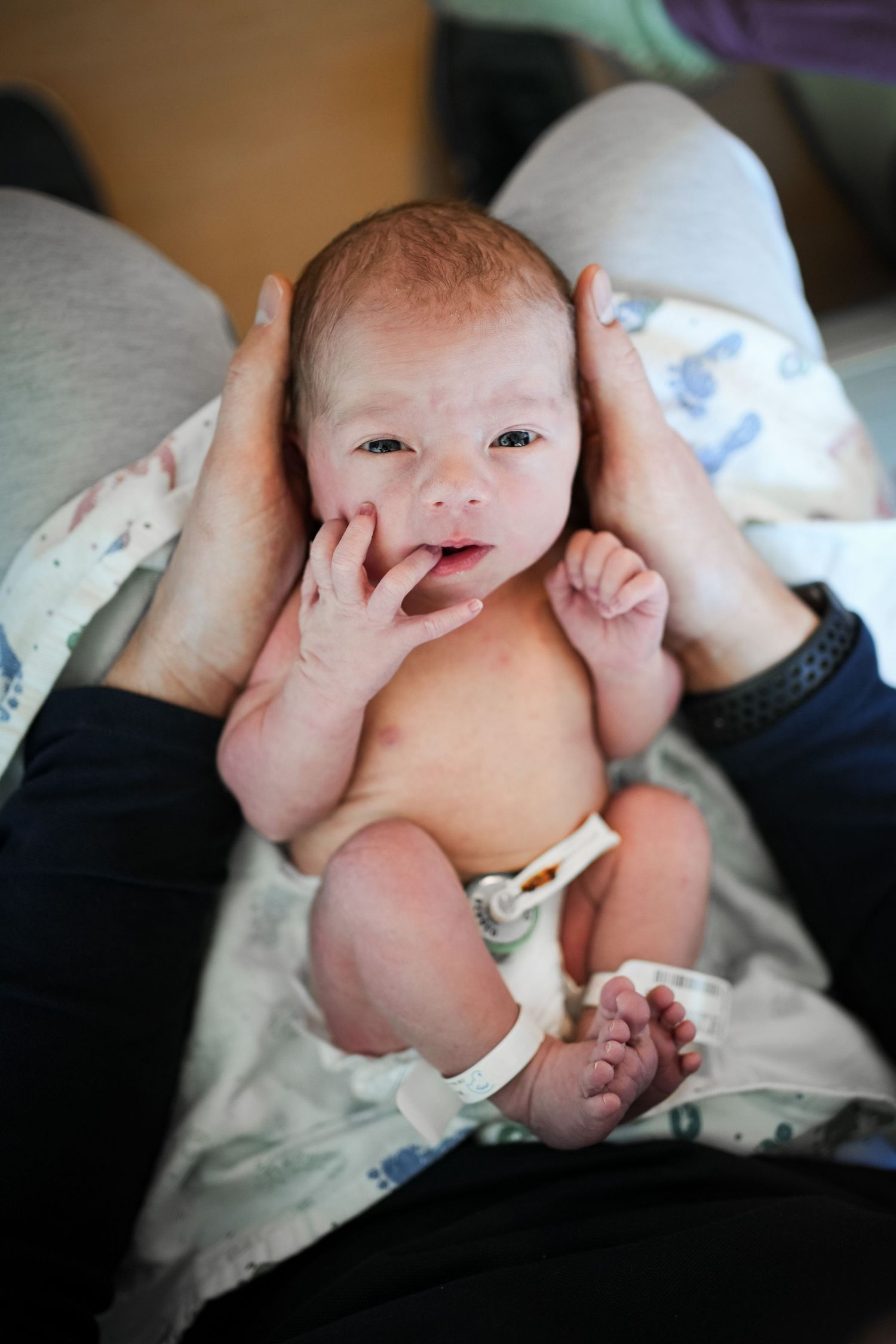 Newborn baby with hospital bracelet lying on adult's lap, eyes open, fingers near mouth.