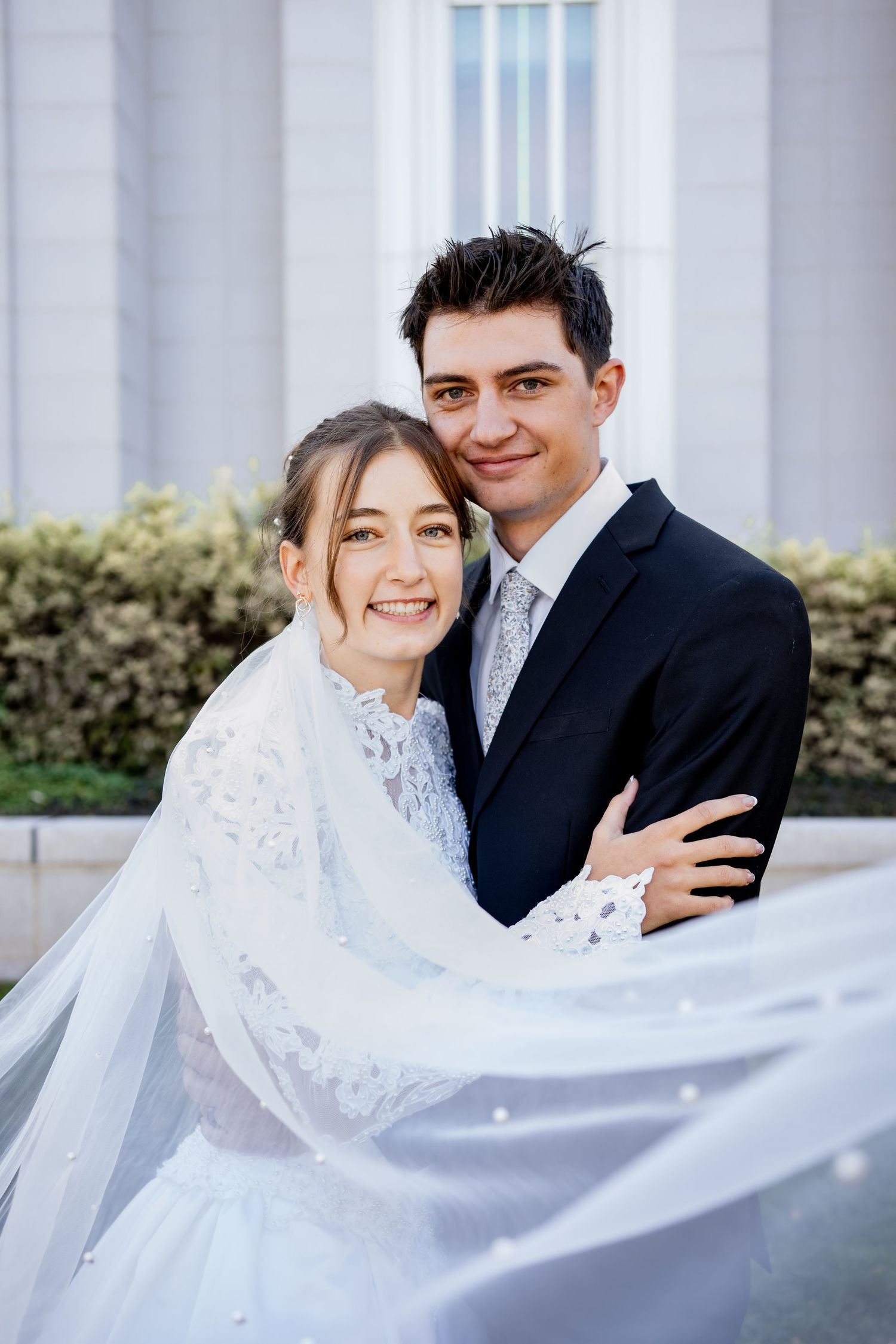 Smiling bride in a lace wedding gown and flowing veil embraces a groom in a navy suit and gray tie outside a light stone building.