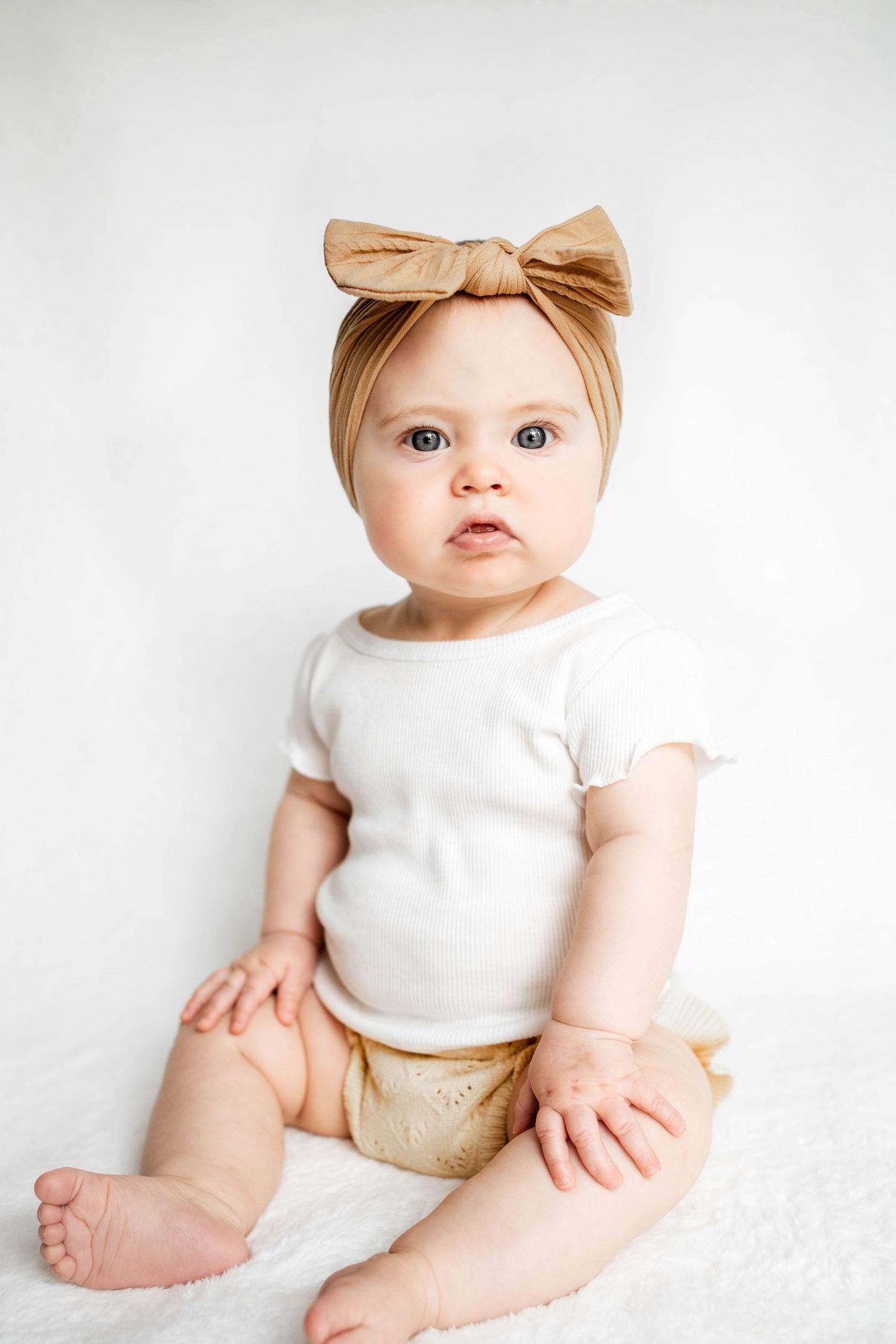 Baby girl in a tan knotted bow headband, white ruffle-sleeve top and tan bloomers sitting on a soft white backdrop.