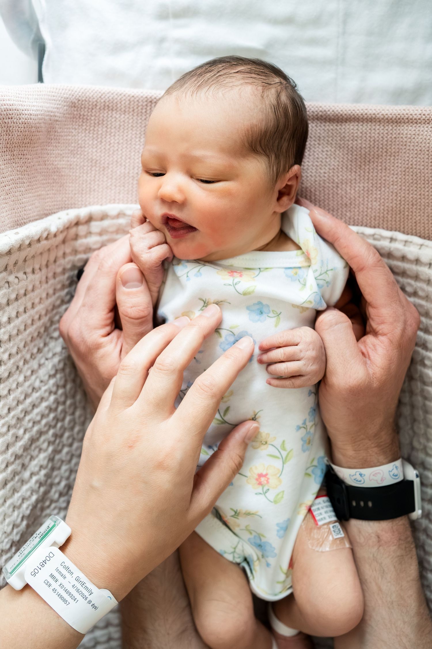 Newborn baby in floral onesie sleeping peacefully in wicker bassinet held by both parents.