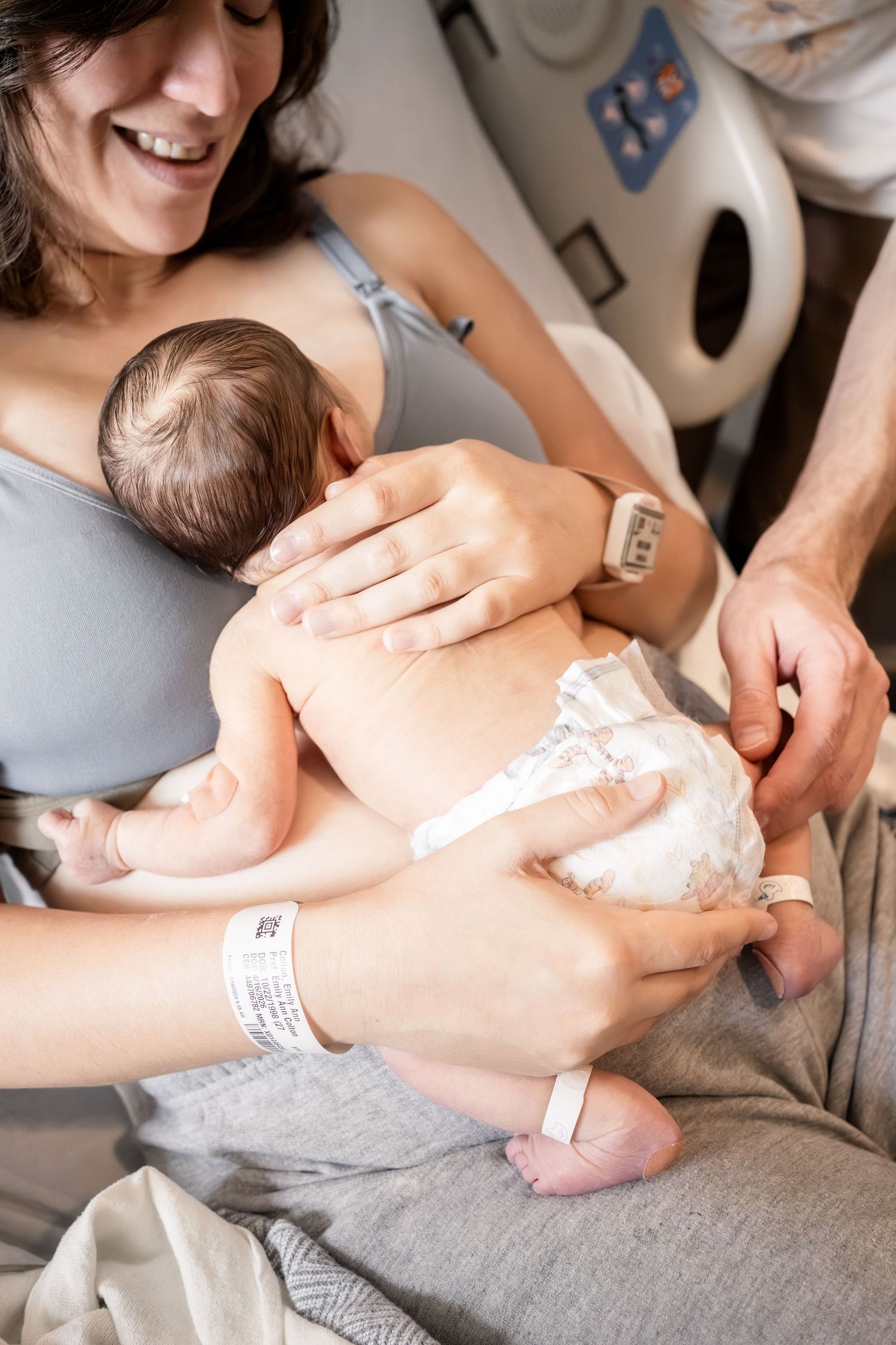 Smiling mother holding newborn baby in hospital bed, with partner's hand visible, both wearing ID wristbands.