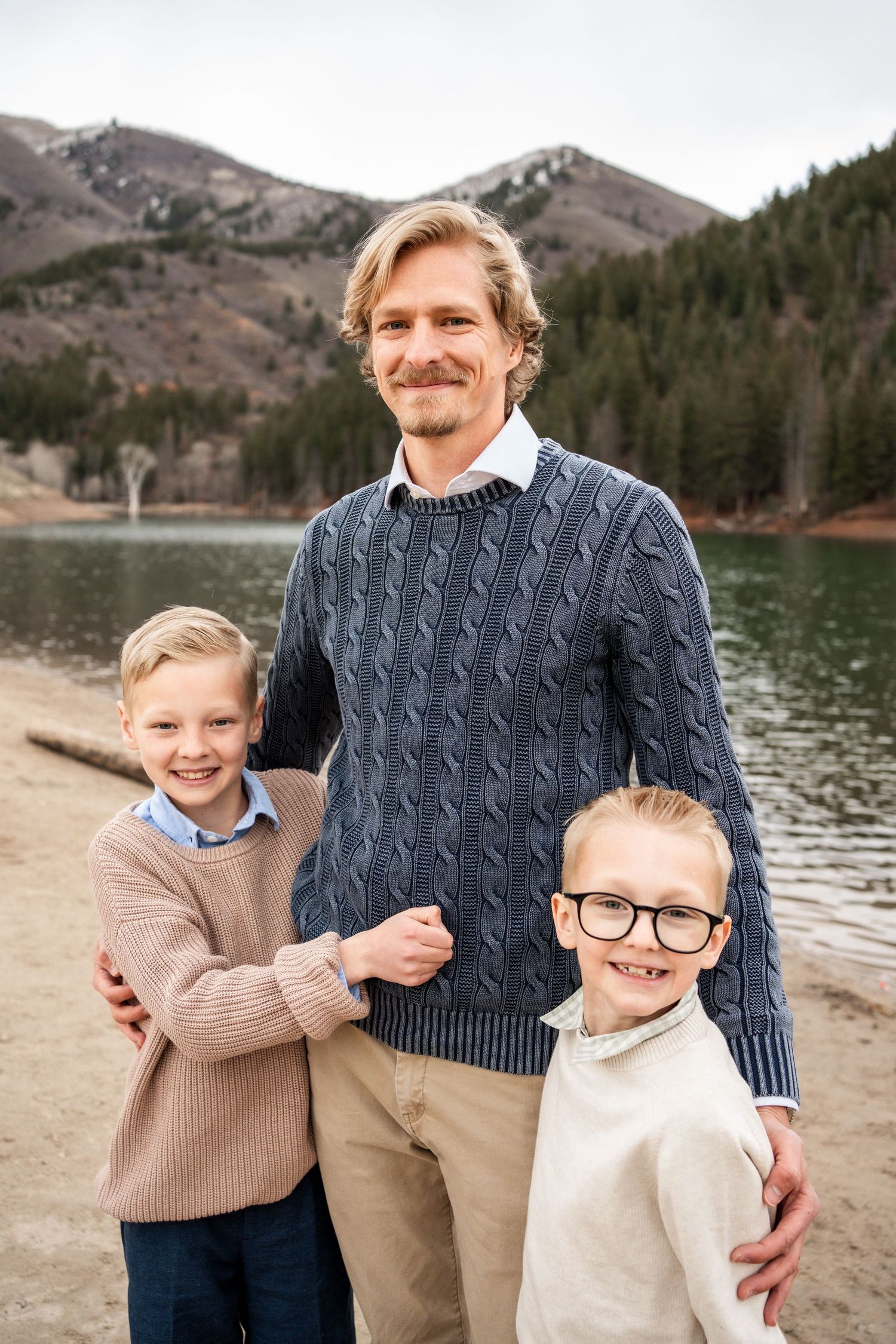 Three people pose together by a mountain lake with evergreen forest in the background.