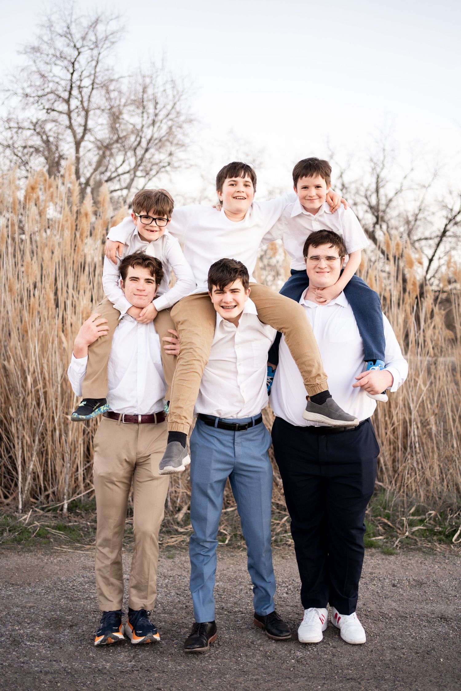 A group of young people in white shirts and casual attire pose together outdoors against tall reeds and bare trees.