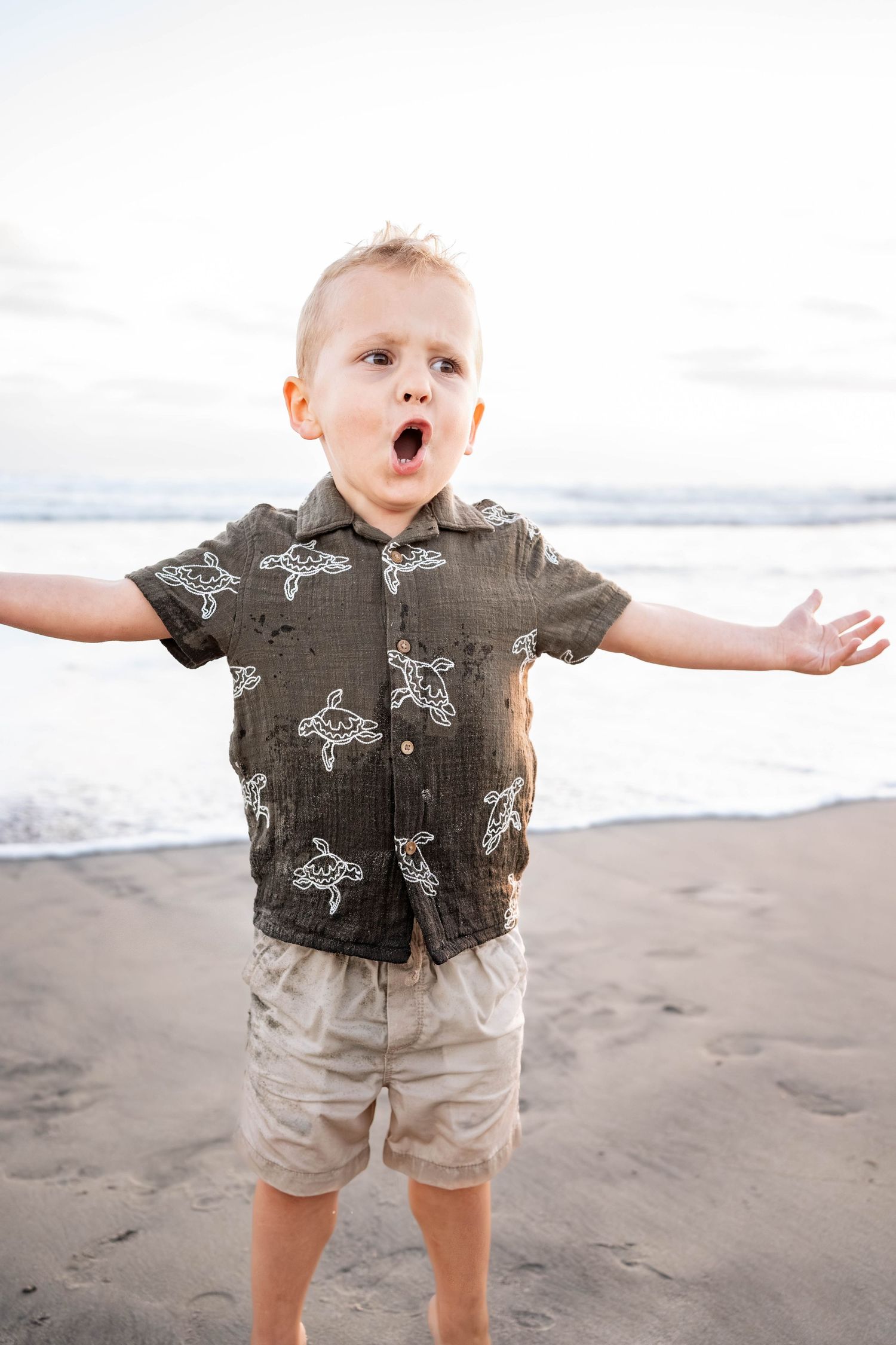 Excited toddler boy with arms outstretched on sandy beach wearing turtle-print shirt and khaki shorts.