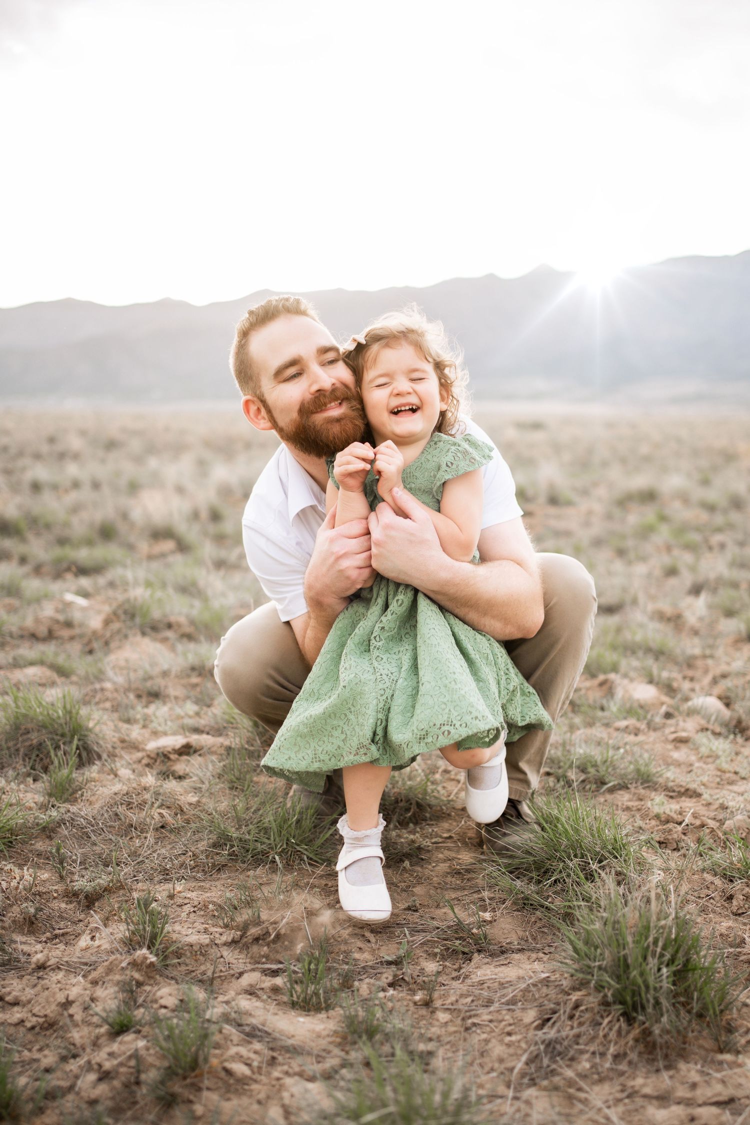 Father crouching and hugging laughing toddler girl in green dress outdoors in a sunlit desert field.