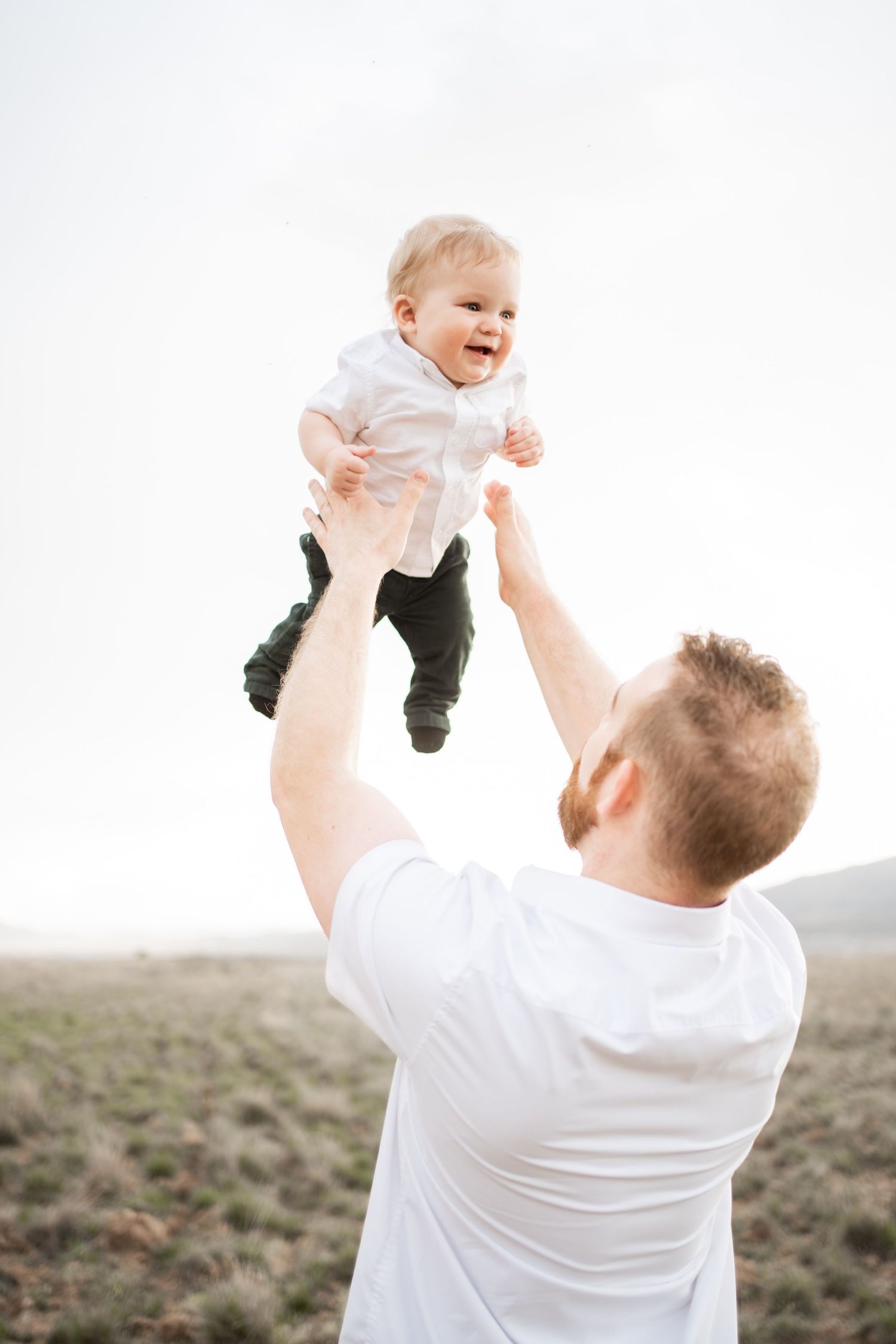 Father in white t-shirt joyfully lifting smiling baby in white shirt and green pants outdoors against a bright sky.