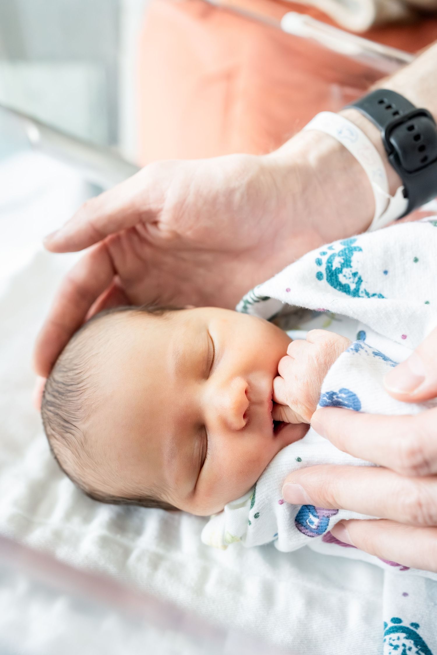 Newborn baby sleeping peacefully in hospital, gently cradled by an adult hand wearing a smartwatch.