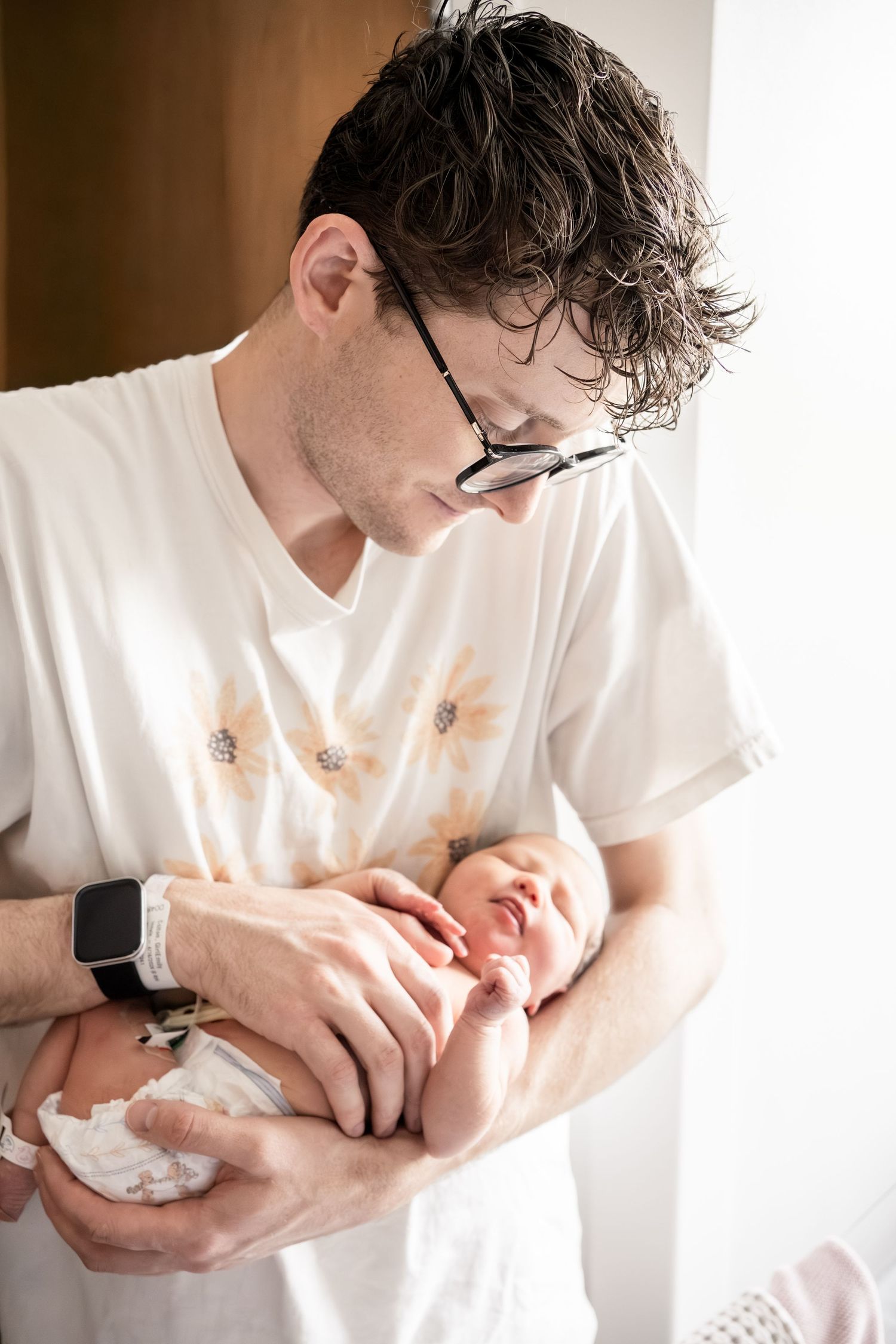 New father in glasses and floral shirt tenderly holding his sleeping newborn baby.