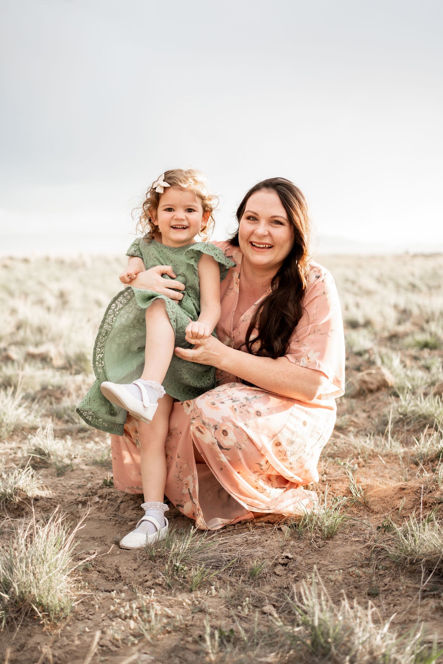 A smiling mother crouches in a field holding her happy toddler daughter, both wearing floral and green dresses outdoors.