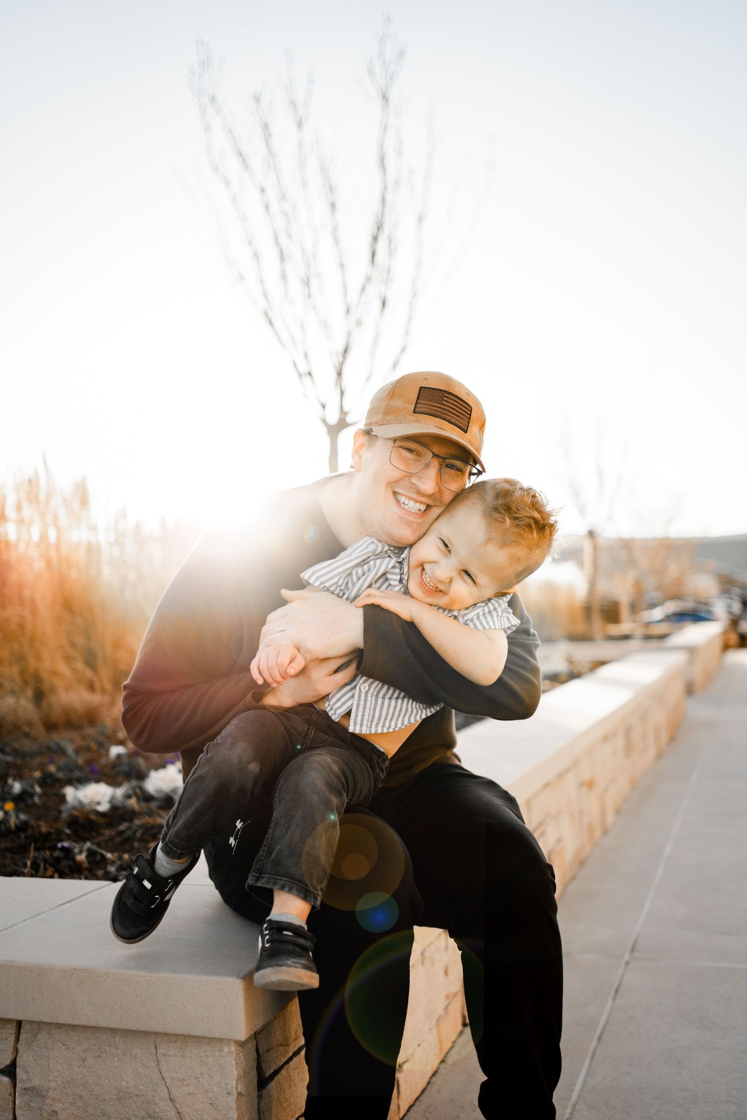 A parent and child share a tender moment sitting together on a concrete ledge during golden hour sunlight.