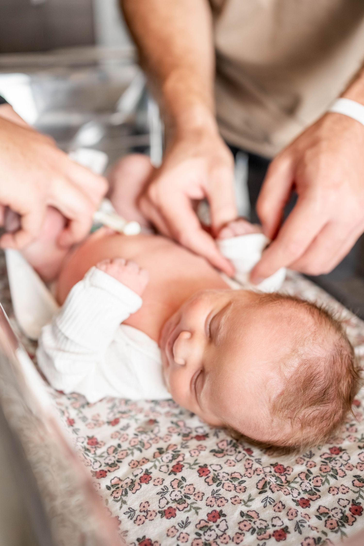 Newborn baby being dressed by parents in hospital nursery, wrapped in white onesie on floral blanket.