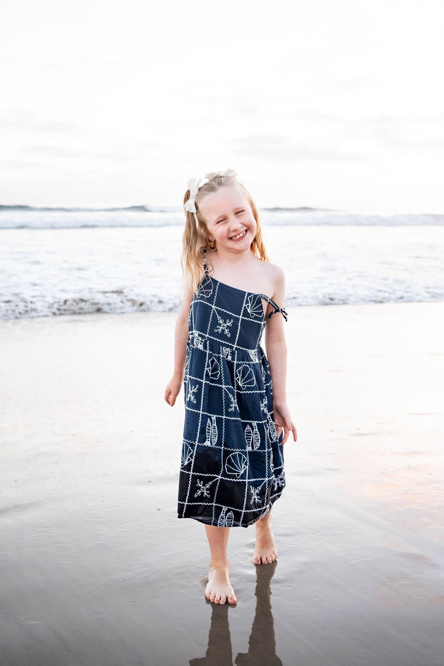 A happy young girl in a navy seashell print sundress stands barefoot on a wet sandy beach, smiling brightly.