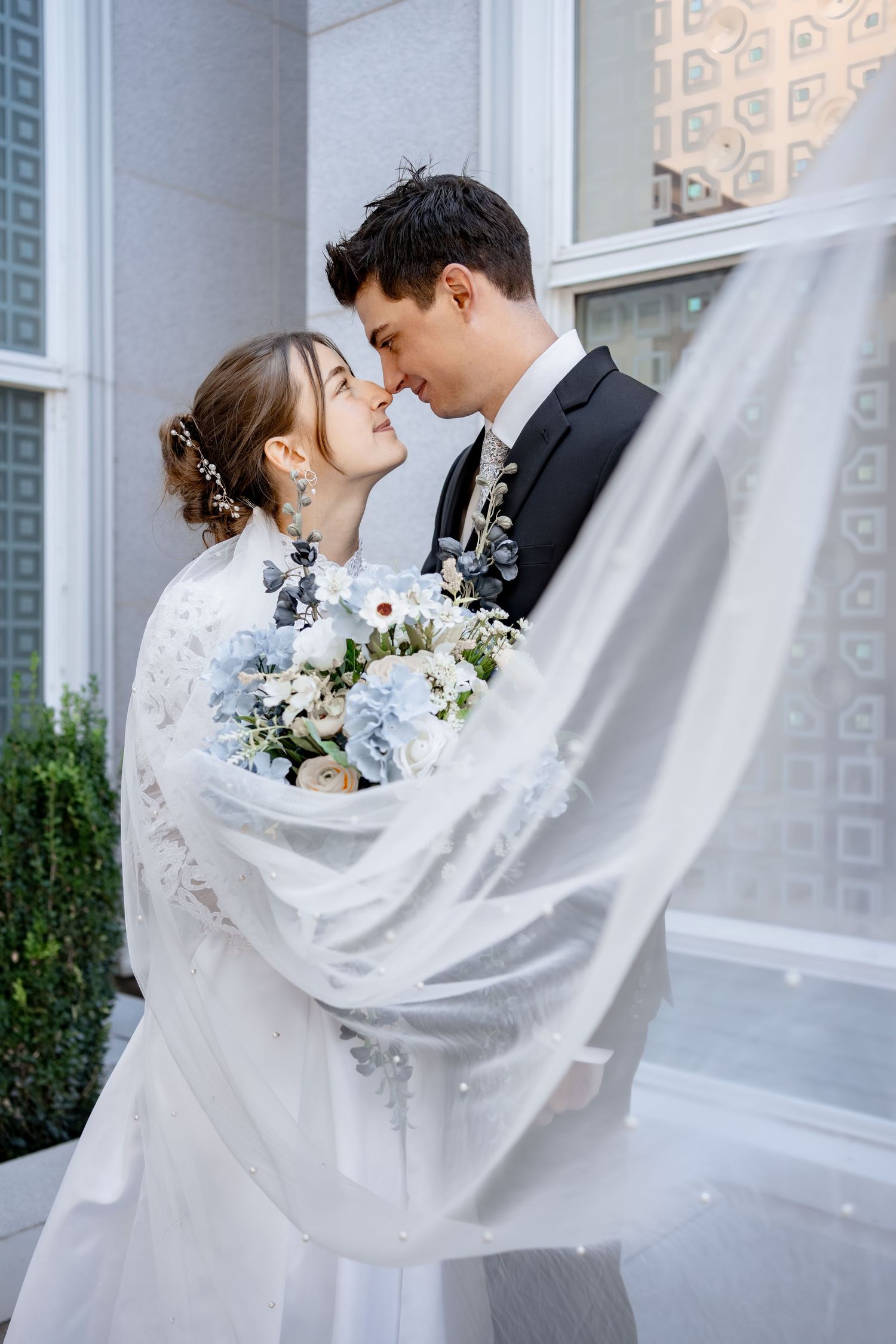 Bride in a lace gown and flowing veil touches noses with her groom while holding a blue and white floral wedding bouquet.