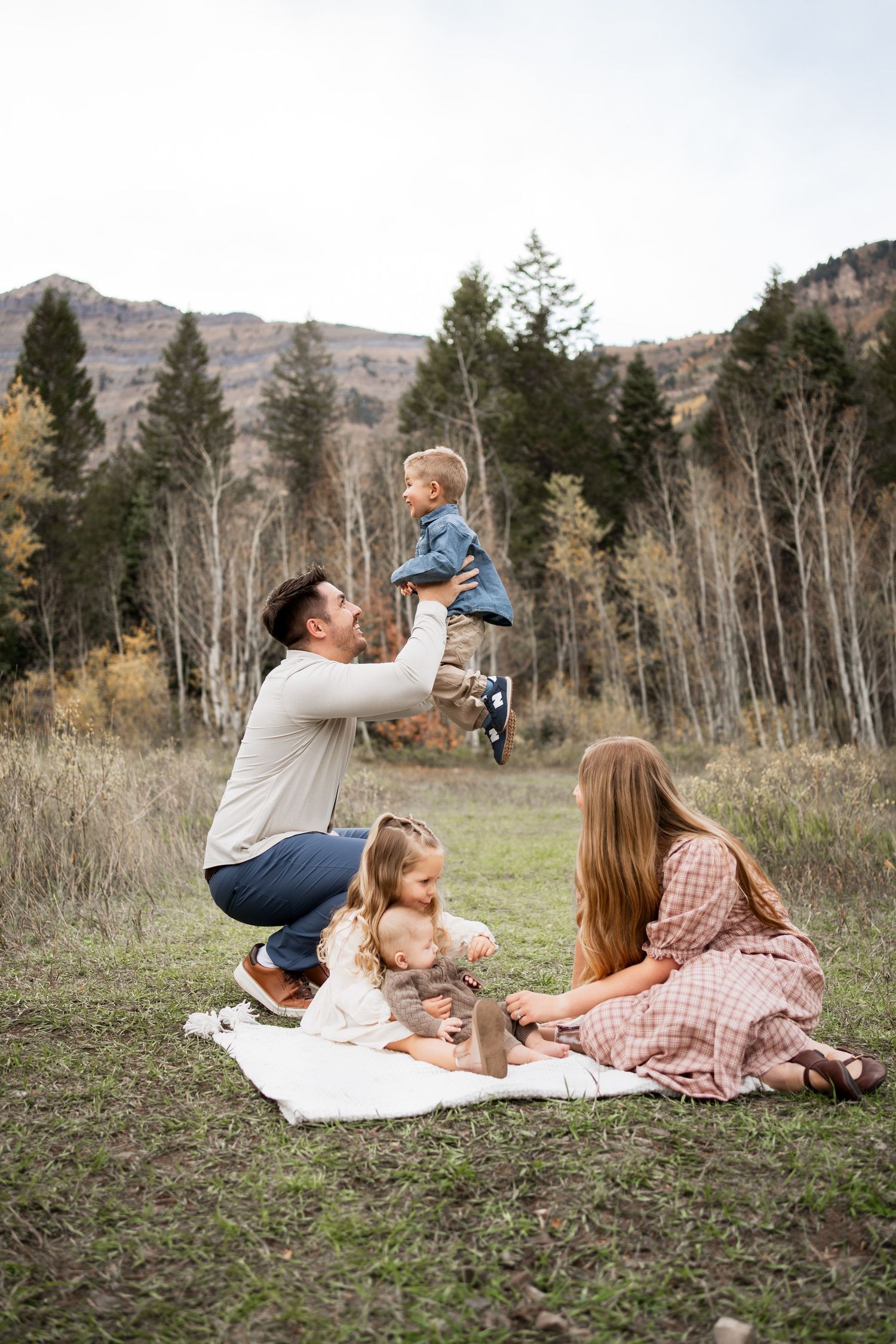 Family of five enjoying an autumn picnic on a blanket in a mountain meadow while a father lifts a laughing toddler into the air.