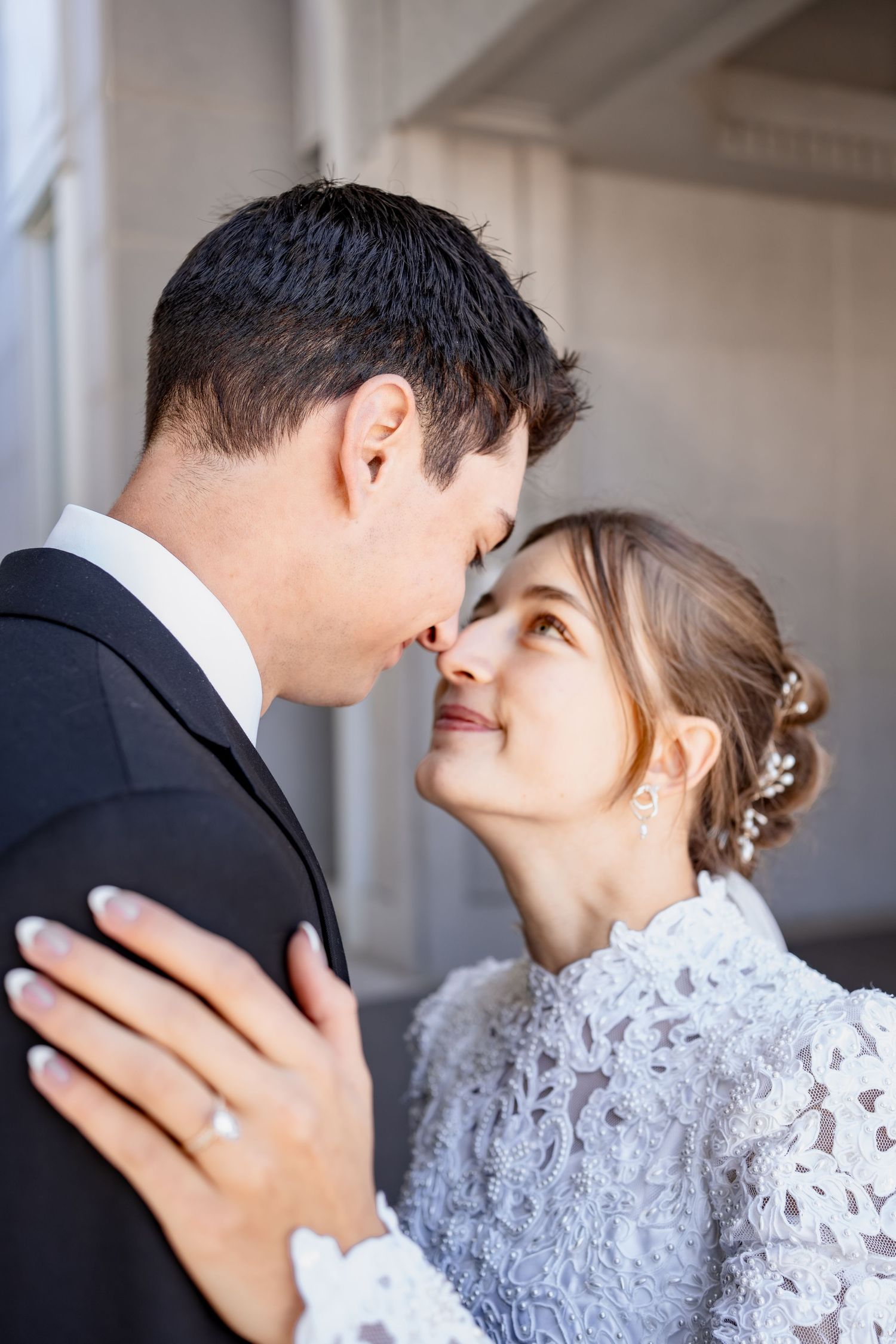 Smiling bride in a white lace dress and floral hair accessories gazes lovingly at a groom in a dark suit during a romantic wedding portrait.
